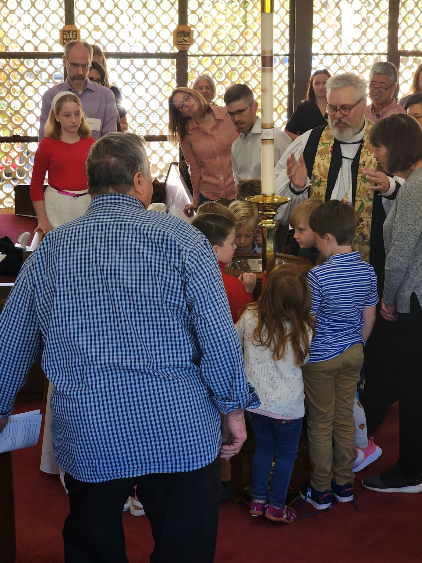 A group of children gather around a clergyman at a church altar. Several adults watch.