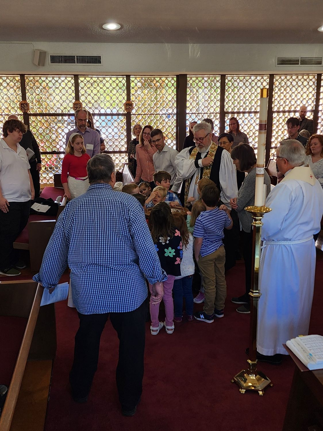 People, including children, in a church. A priest and others bless them. Sunlight streams through stained glass windows.