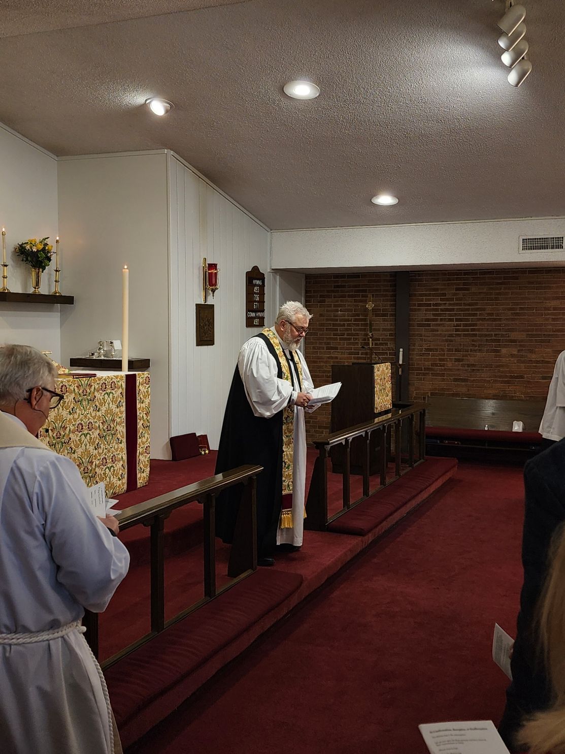 Man in clerical robes reads at a church service; red carpet, brick wall, other people present.