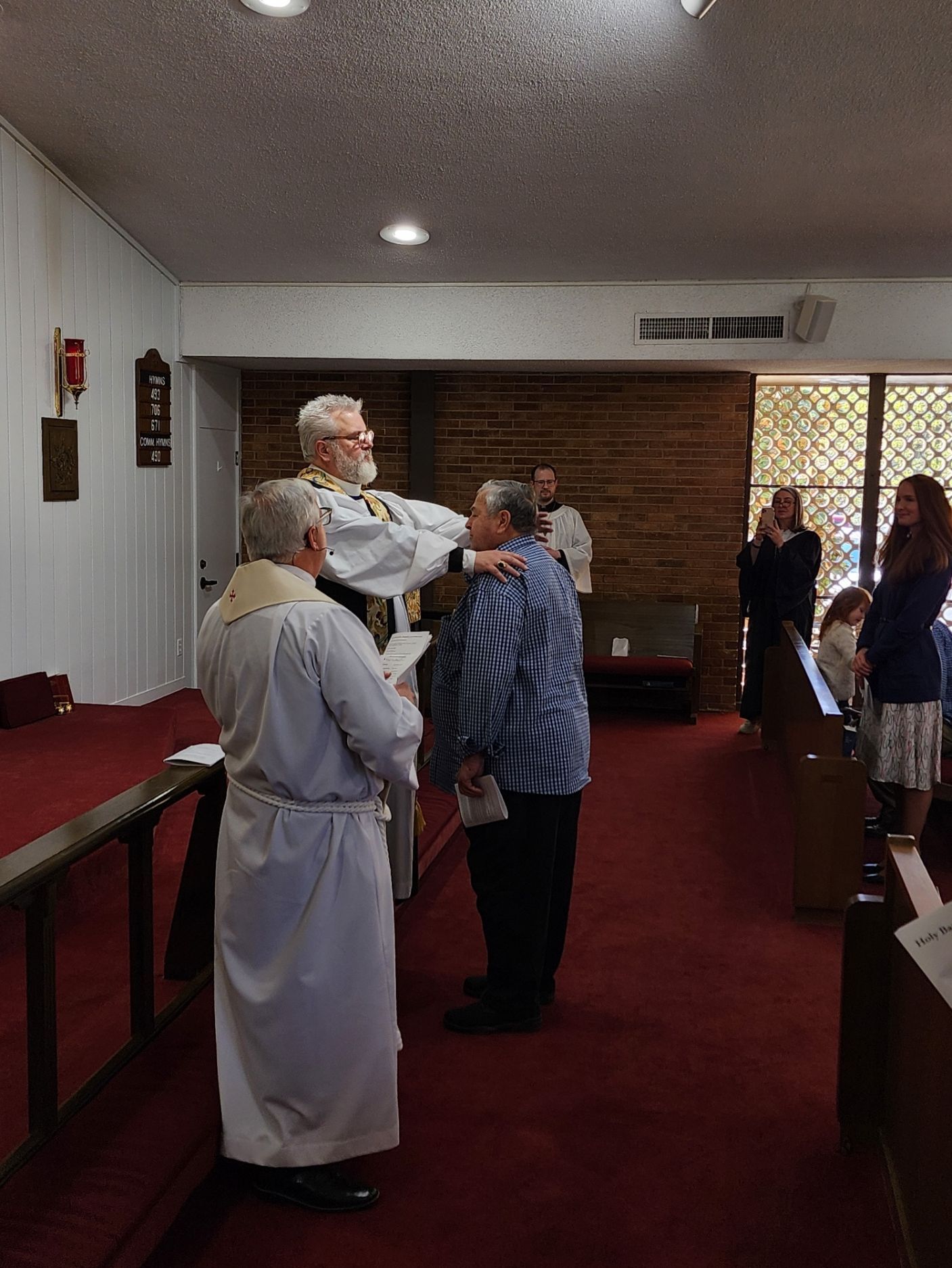 Clergy laying hands on a man's head in a church. Other people watch, some smiling. Red carpet, stained glass.