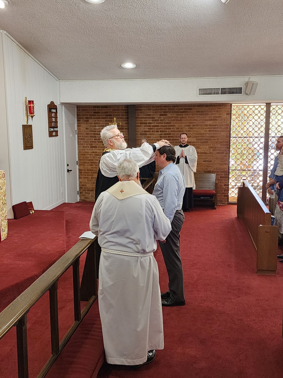 Clergy member anoints a person's forehead in a church with red carpet. Another person stands by.