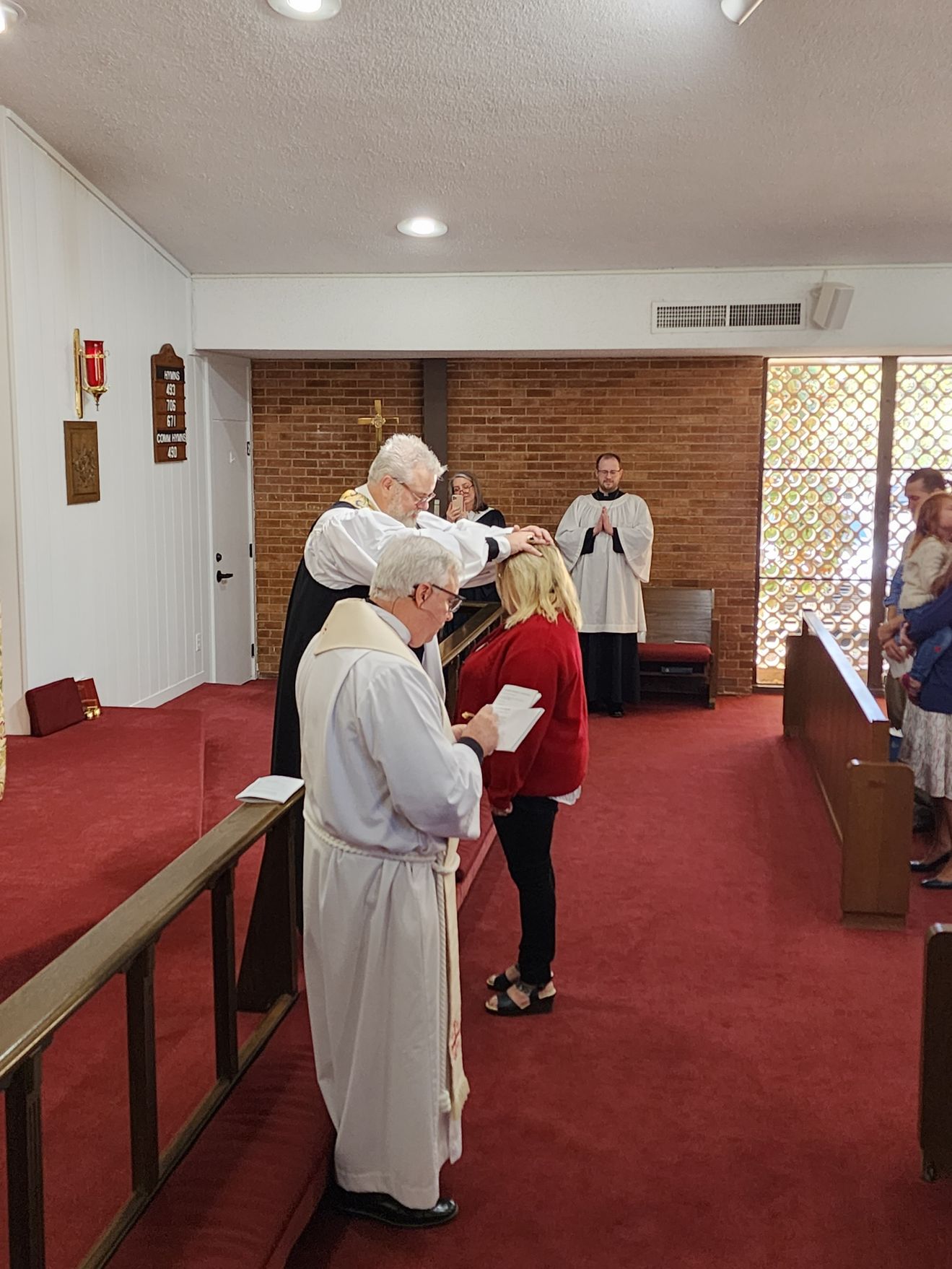 Clergy members place hands on a woman's head during a church service. Red carpet, brick wall, and other attendees visible.