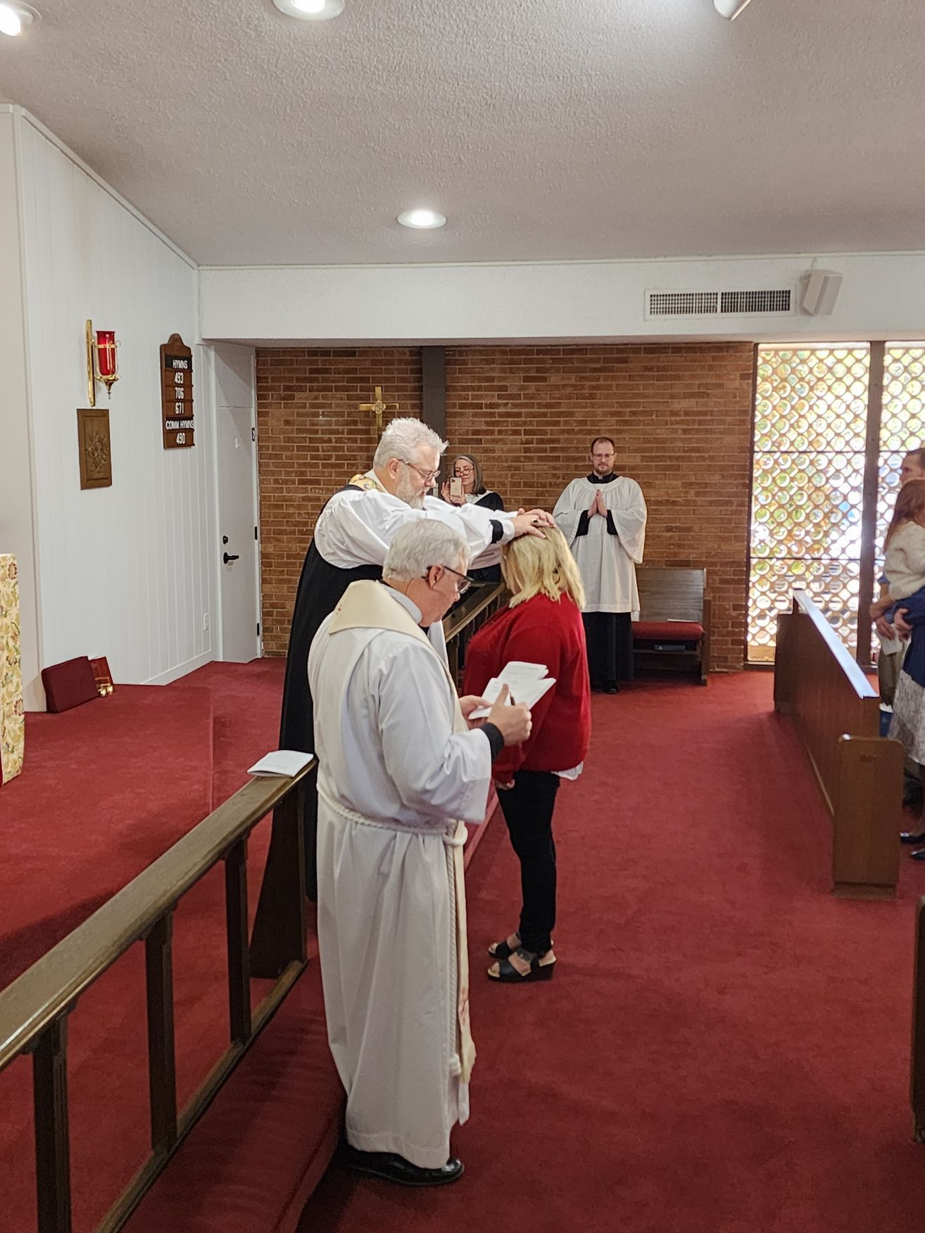 Clergy members anoint a person's head during a church service. Brick wall backdrop.