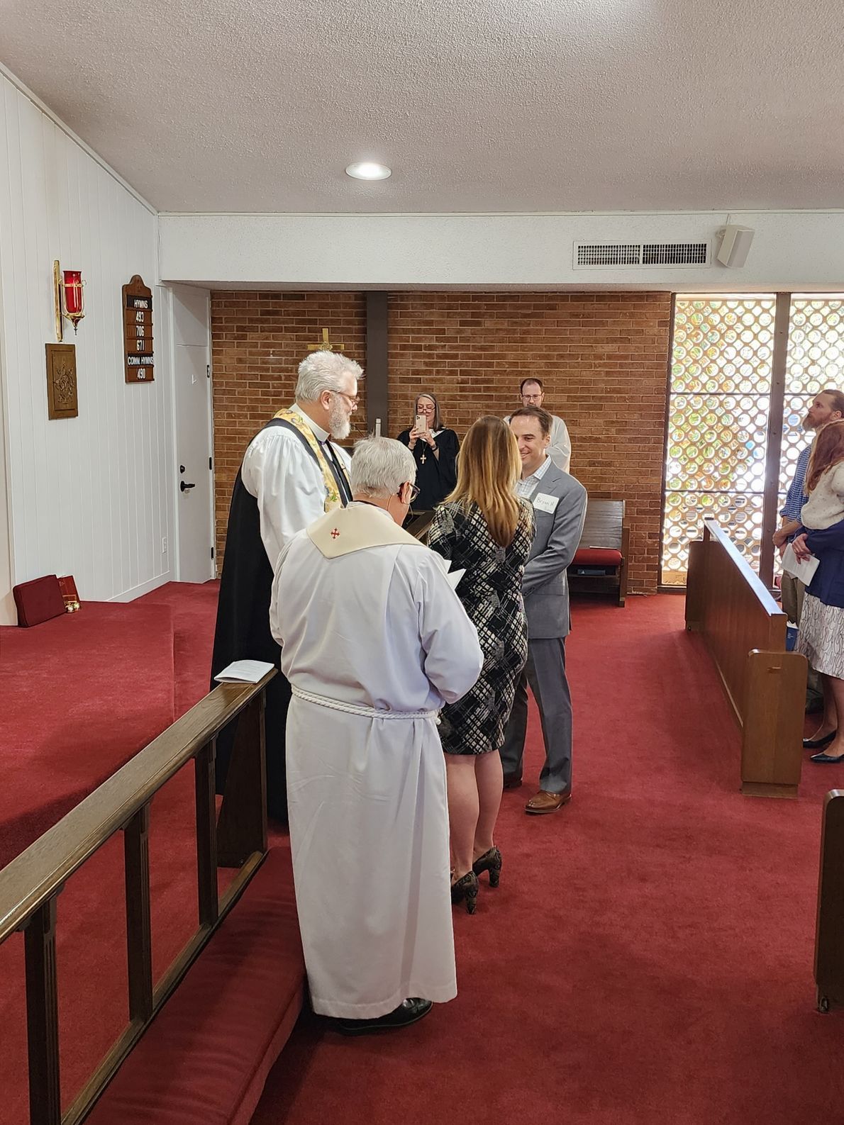 Wedding ceremony in a church: a couple stands before clergy; red carpet, brick wall, and stained glass windows.