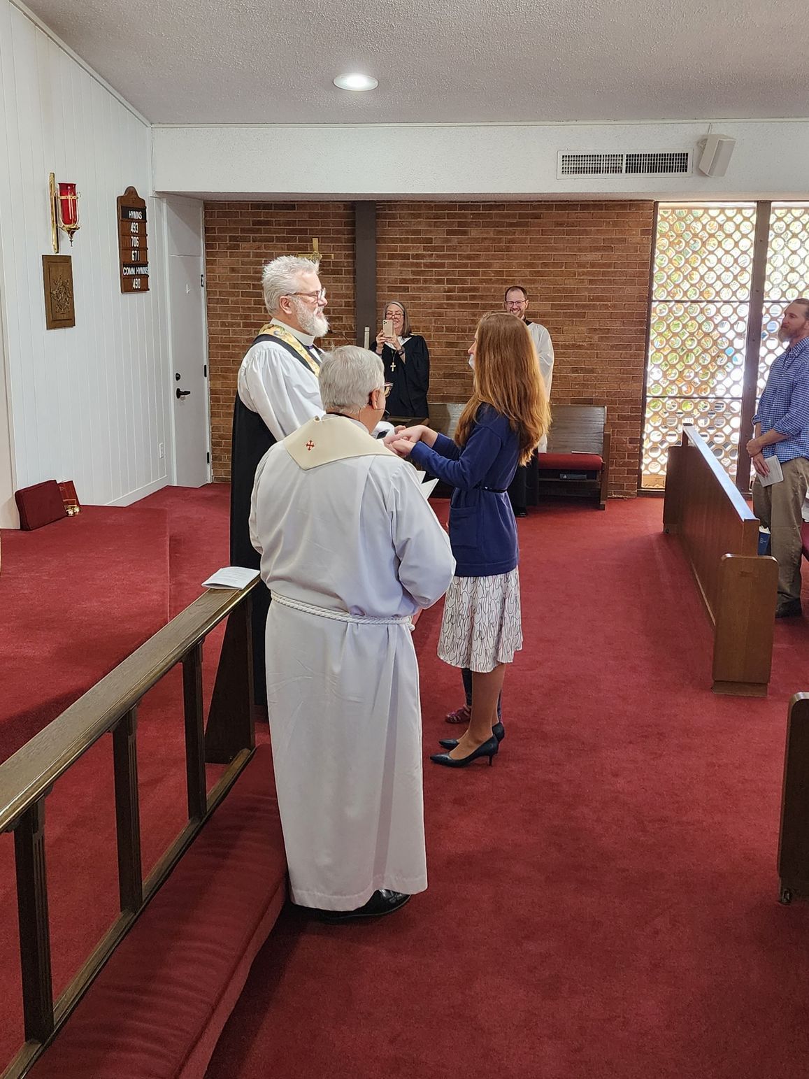 A church ceremony: a woman with a minister in a white robe, another man in a suit, and guests.