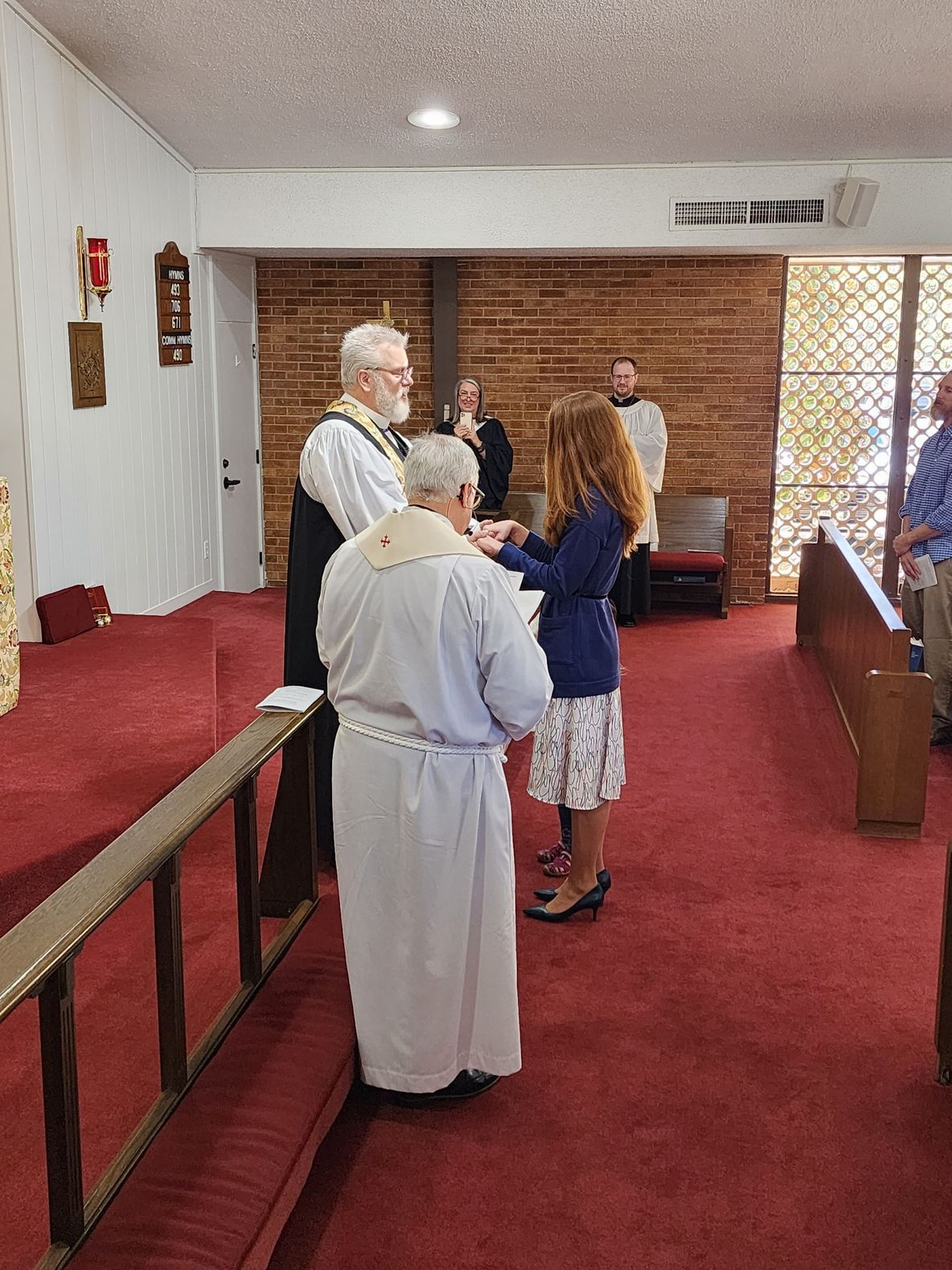 A person receiving a religious rite. Others watch in a church with red carpet.