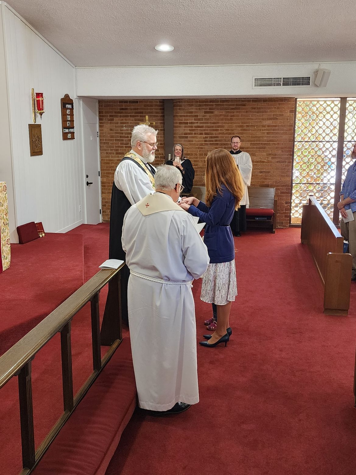 Church service: Clergy in white robes place hands on a person’s head. Brick wall backdrop, red carpet, wooden pews.