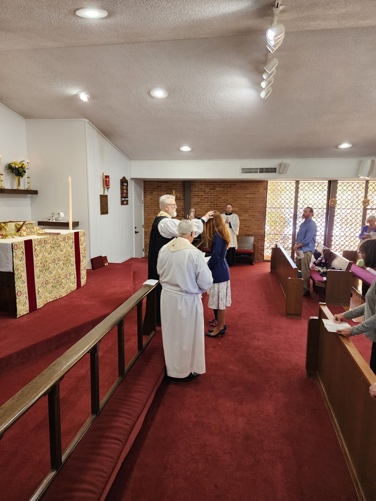 Church service with clergy laying hands on a woman; other attendees observe in a room with a red carpet.