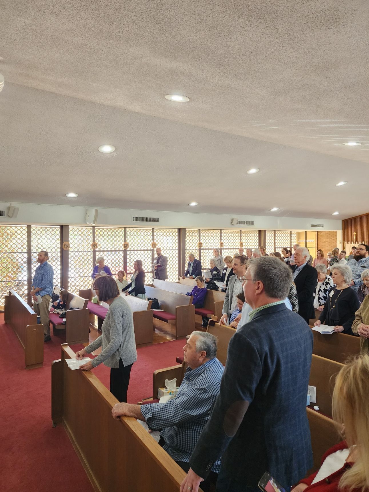 People standing and singing in a church, facing a raised platform. Red carpet, wooden pews, and large windows.