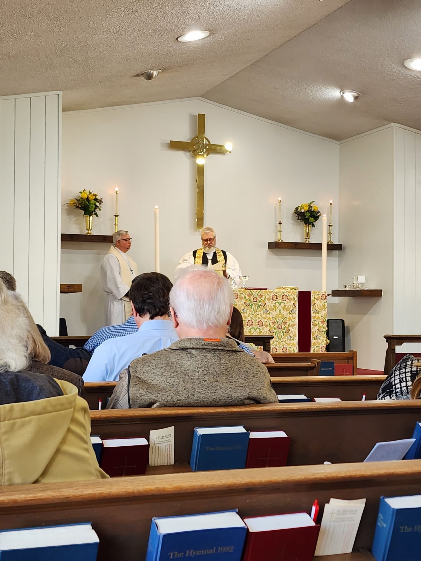 Church service in progress. Priest at altar with assistant. People in pews; gold cross and candles.