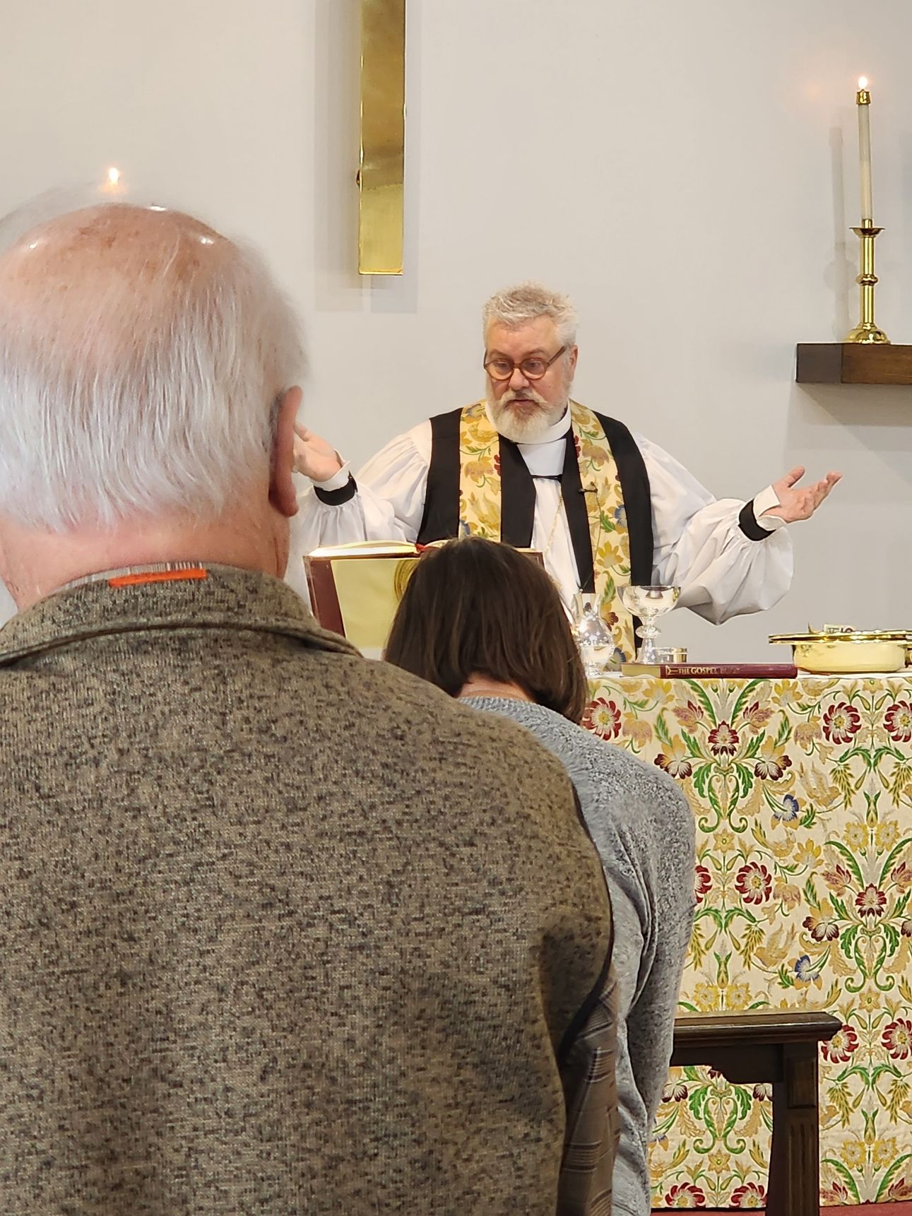 Clergy in white and black vestments, with hands raised, leading a church service. People at altar.