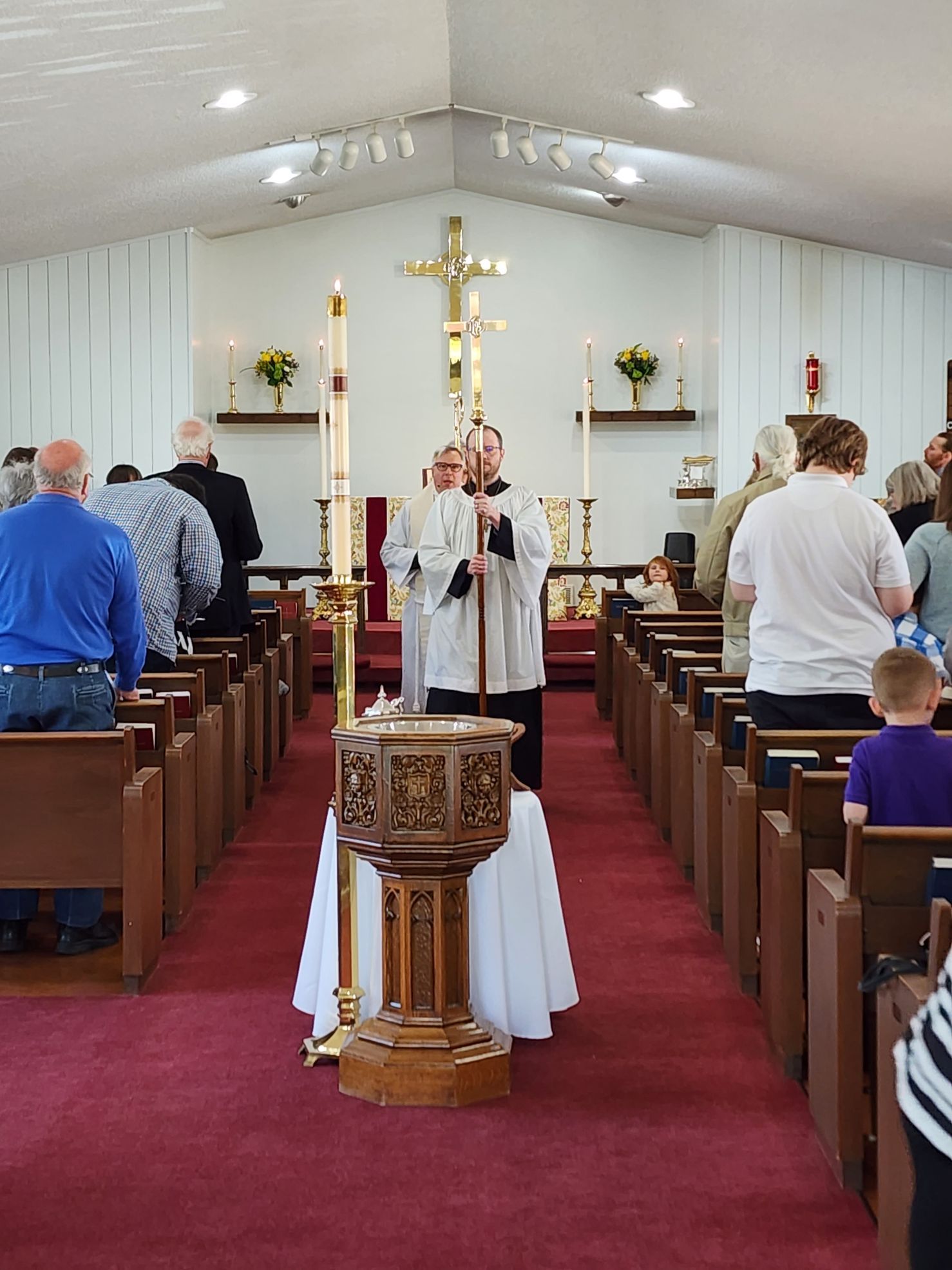 Clergy holding staff, standing before a baptismal font in a church, facing the congregation.