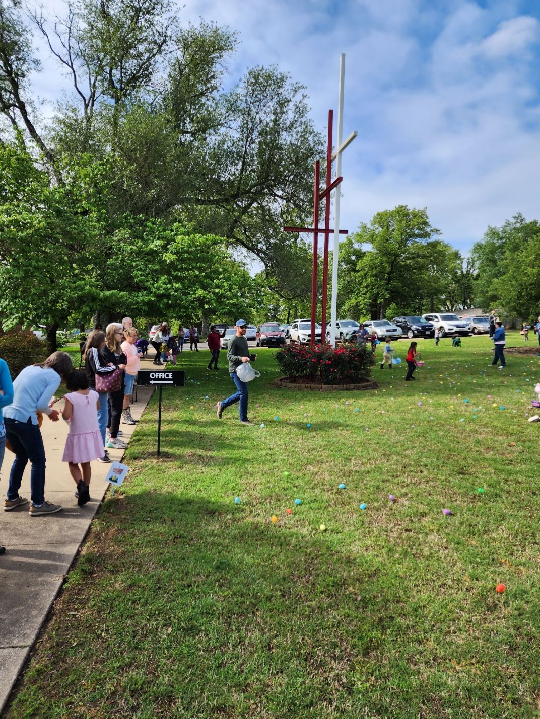 People gather outdoors for an Easter egg hunt on a grassy lawn; a cross stands in the background.