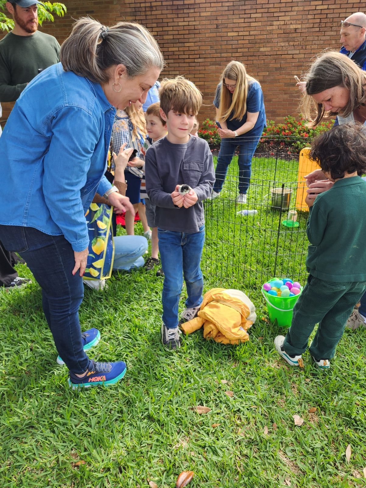 People participating in an Easter egg hunt in a grassy yard. A woman assists a young boy; others search.