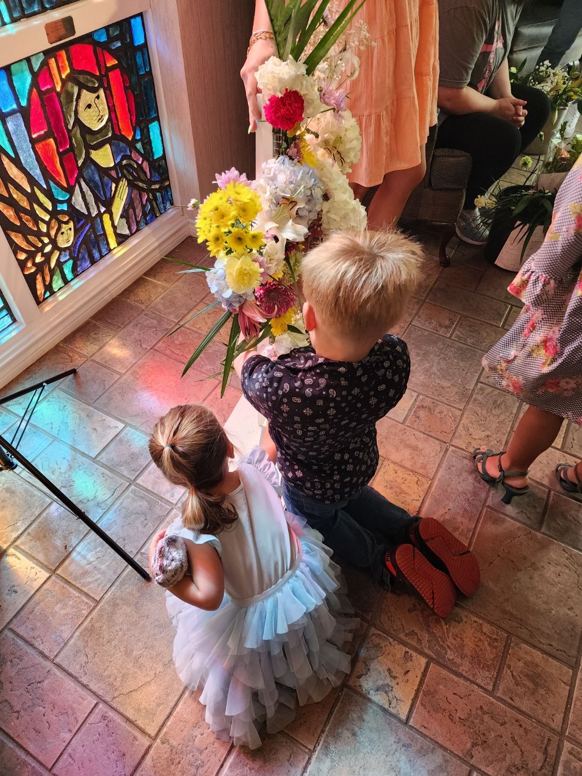Two children kneeling before a flower arrangement; stained-glass window in background.