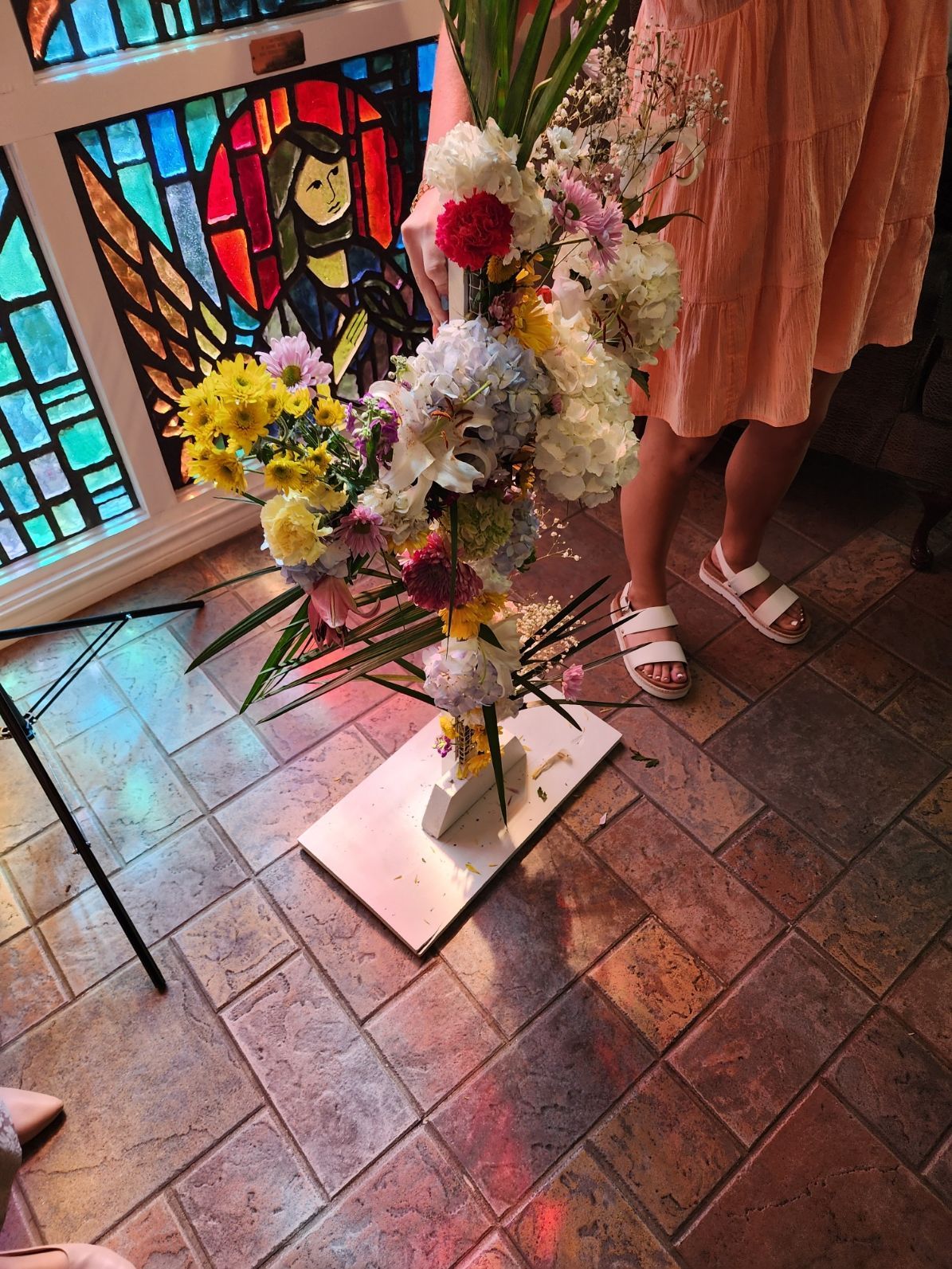 Bouquet of colorful flowers on a pedestal in front of a stained-glass window, person in pink dress and sandals.
