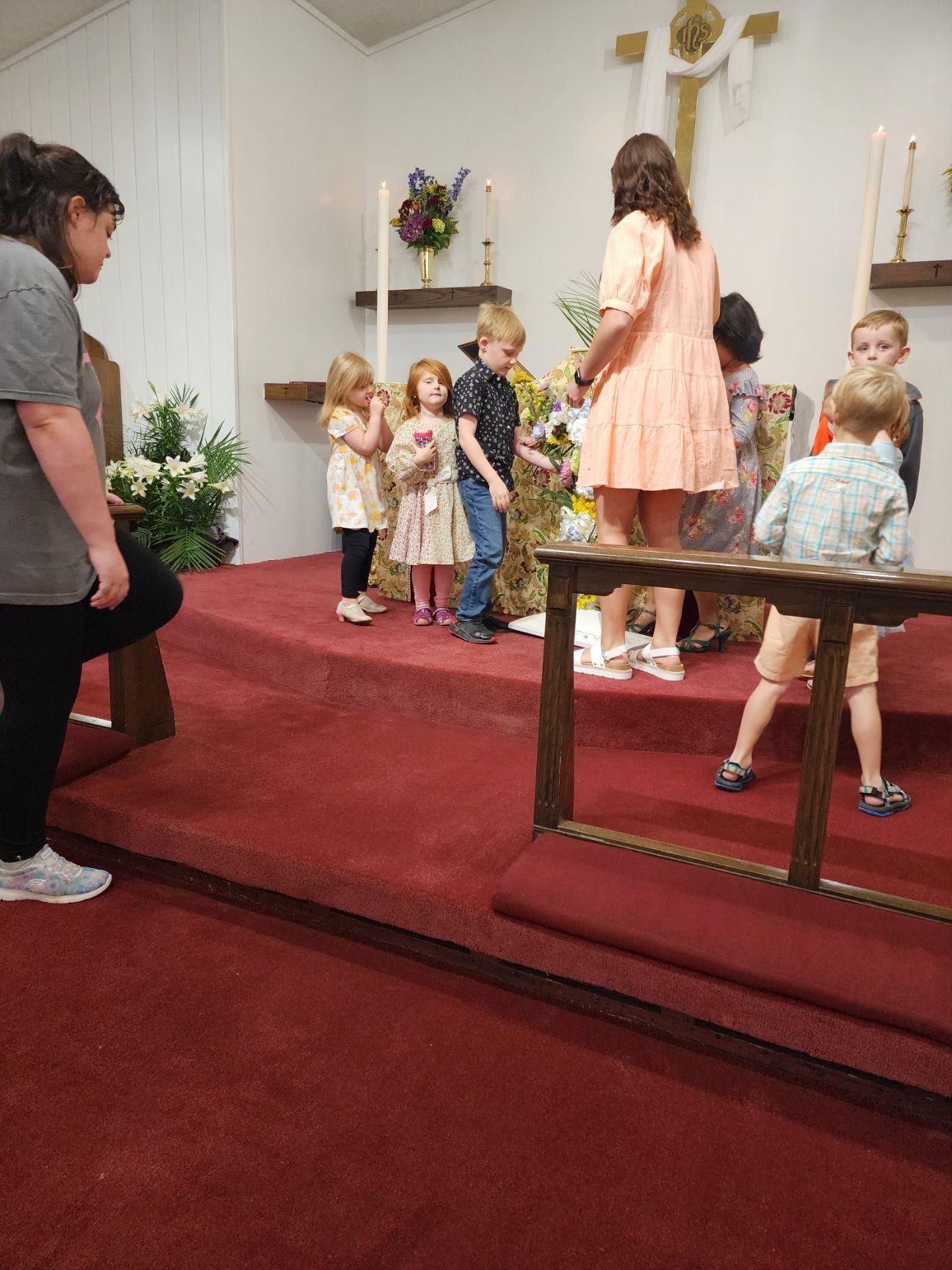 Children and adults on a church stage, with flowers and a cross, likely for a ceremony.