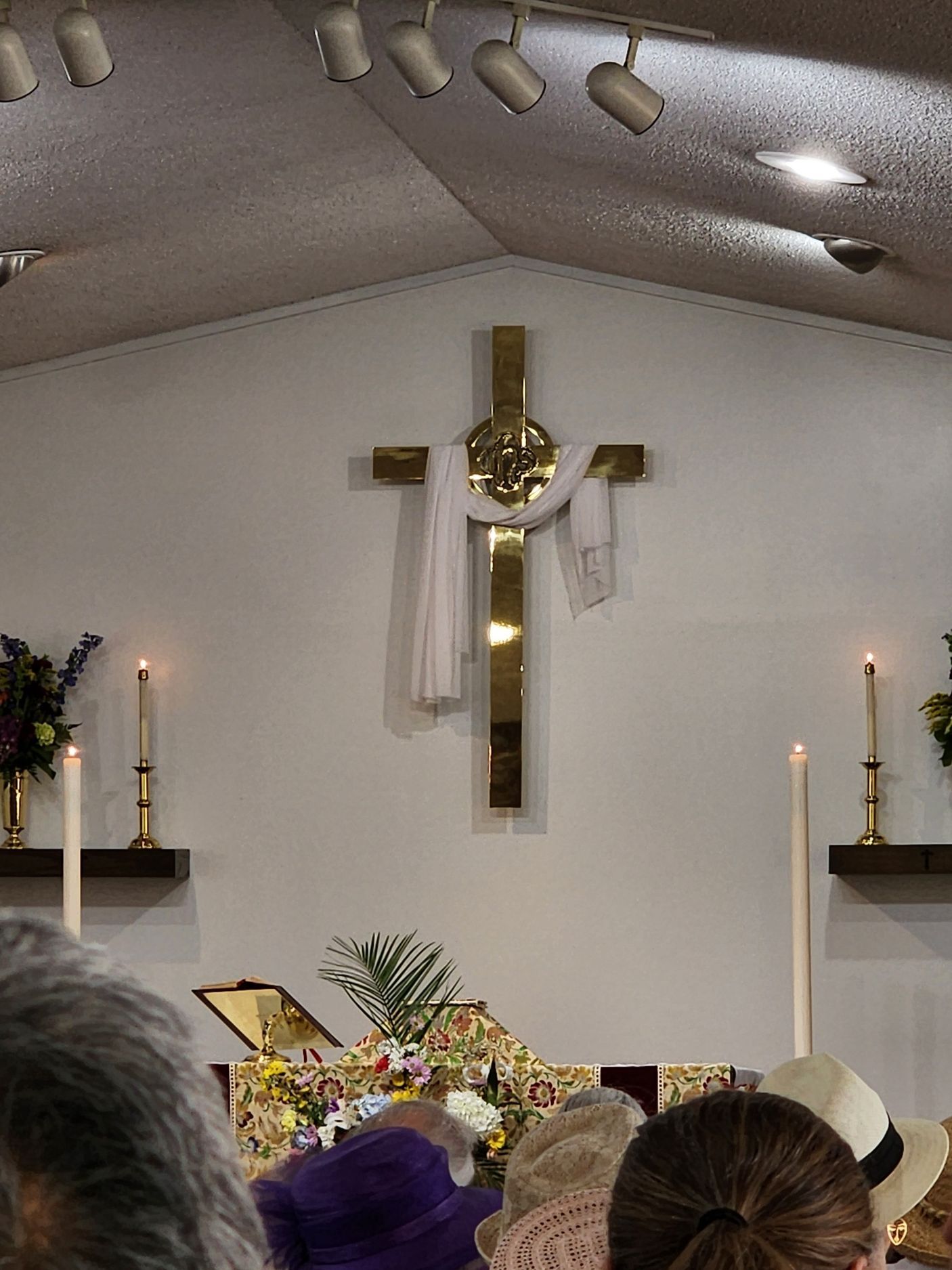 Gold cross with white cloth hanging, on church wall; people and altar in foreground.