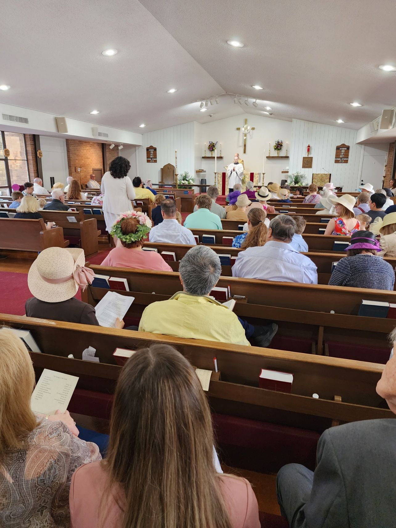 People in pews at church service. Priest at the altar with cross, other figures in front of congregation.