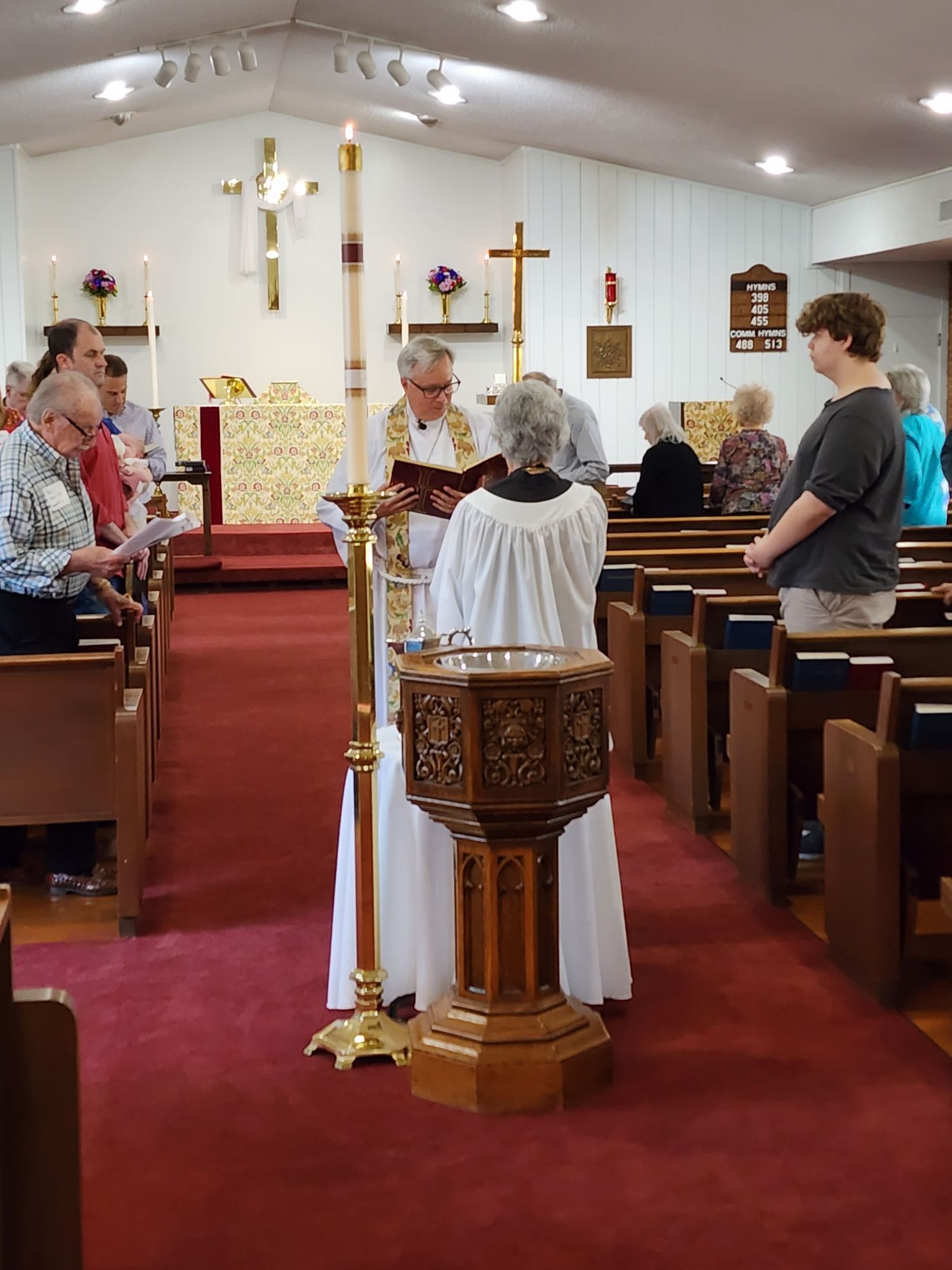 Church service: priest with book, baptismal font, people in pews, tall white candle, red carpet, cross on wall.