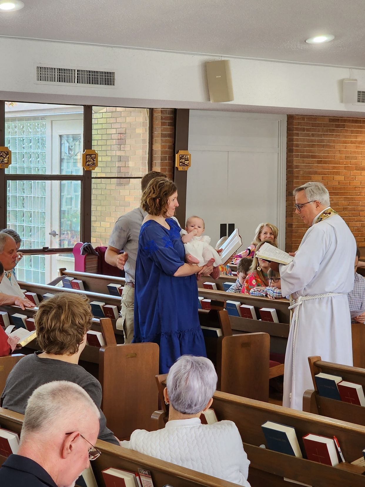 Baptism ceremony in a church; woman holds a baby, priest reads. Others seated in pews.