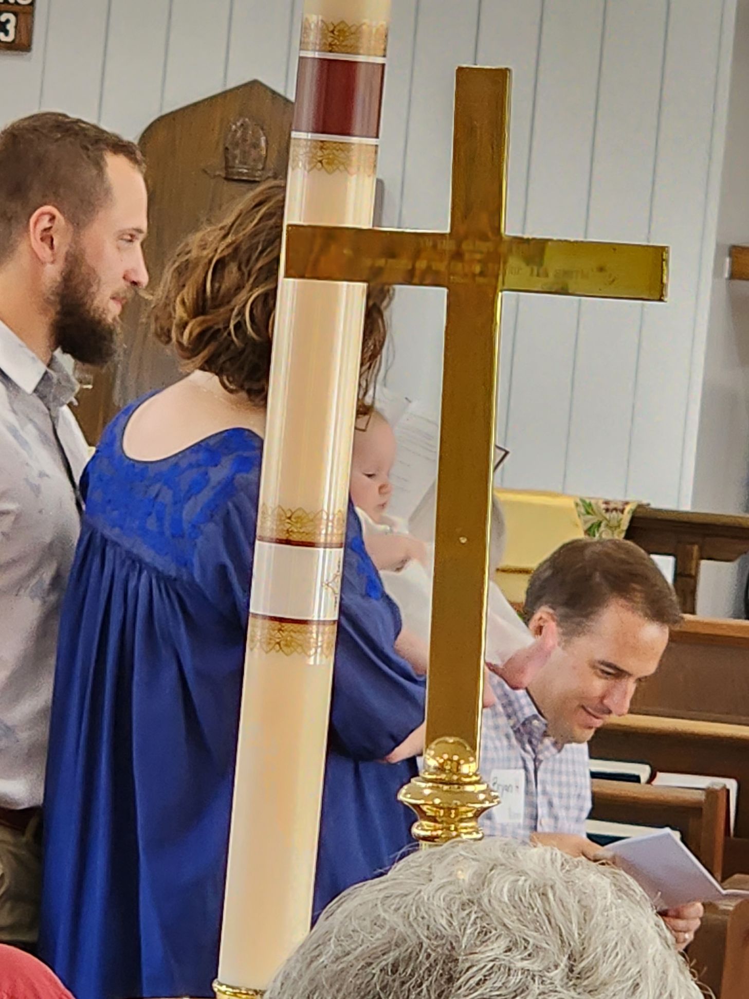 Family at a christening; mother in blue holding baby, man at the side, gold cross in foreground, church interior.