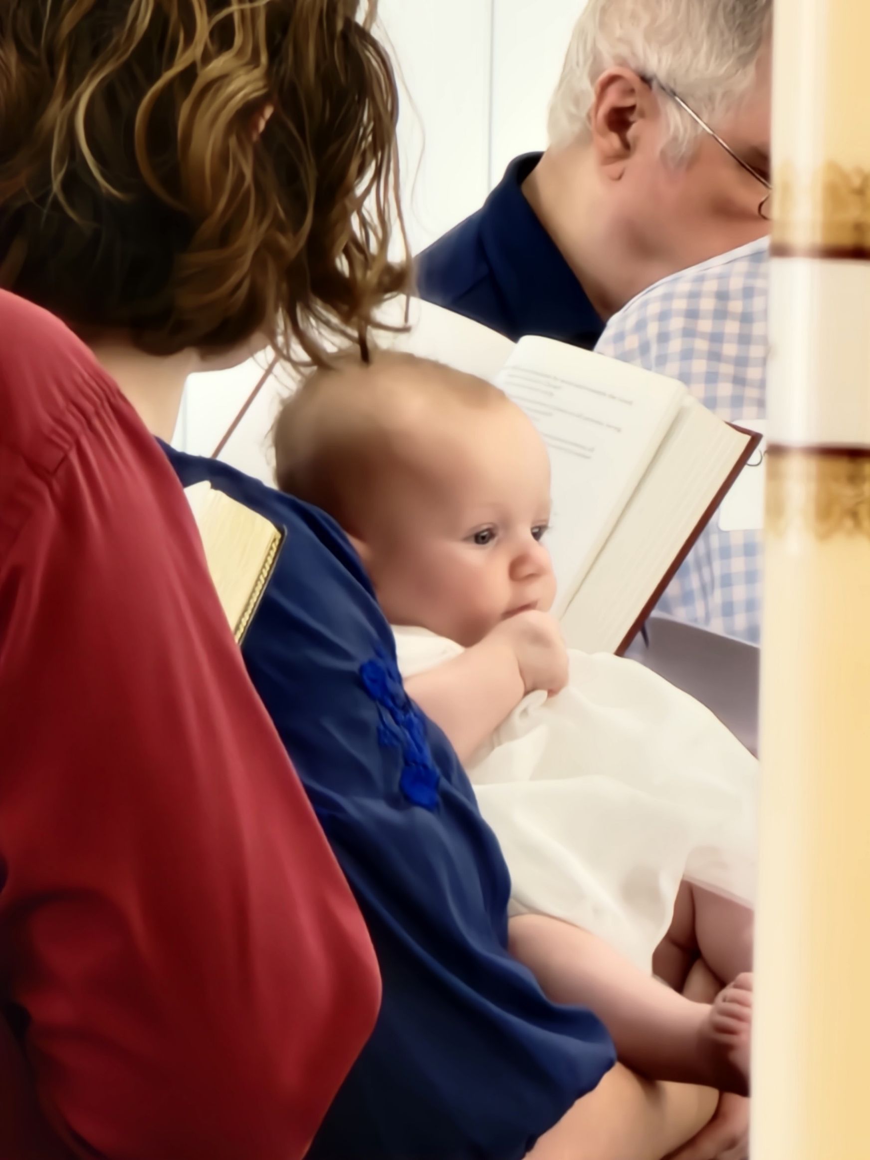 Baby in white dress and blue shirt held by person in red shirt, near a person reading a book.