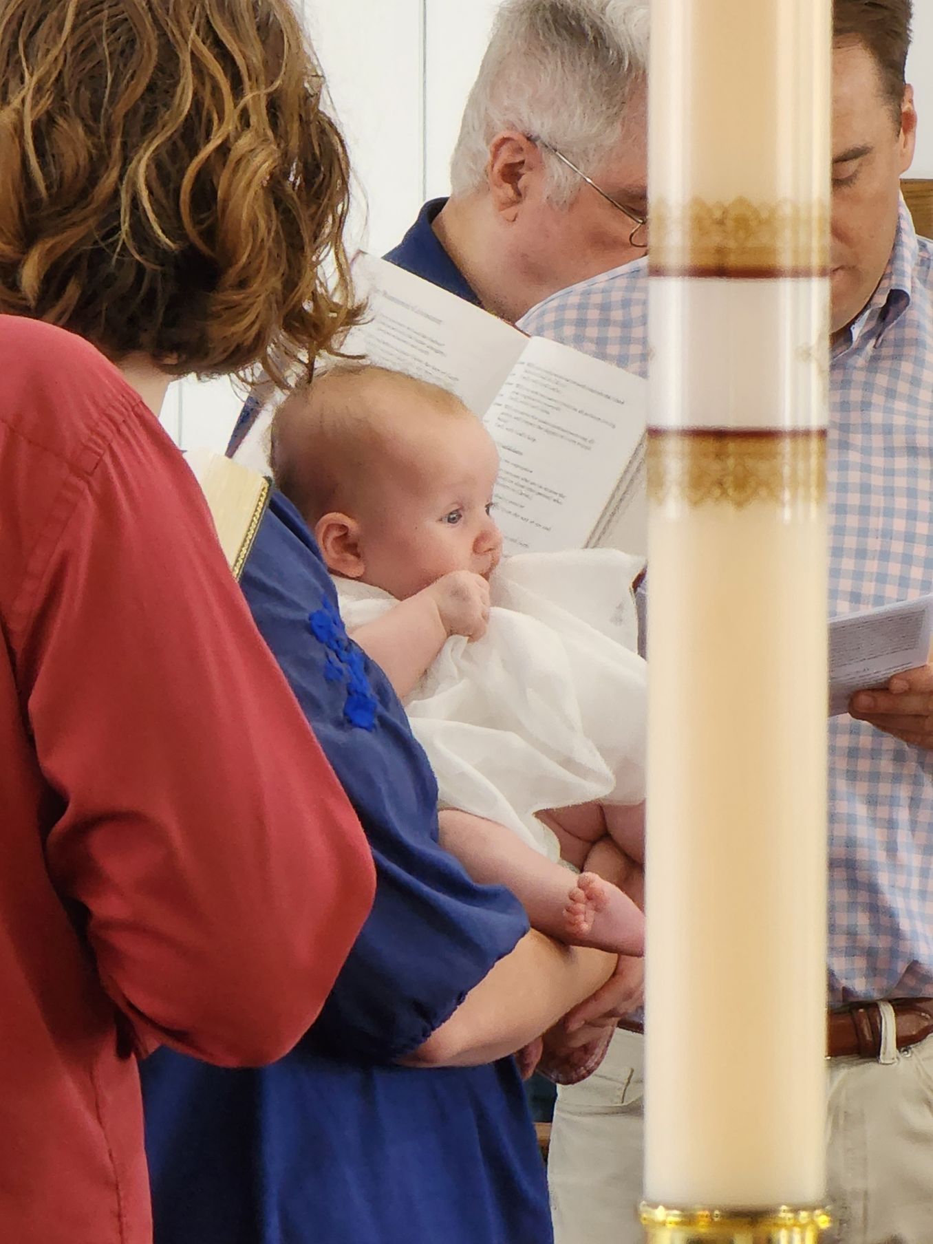Baby in white dress held by person in blue shirt during a baptism ceremony.