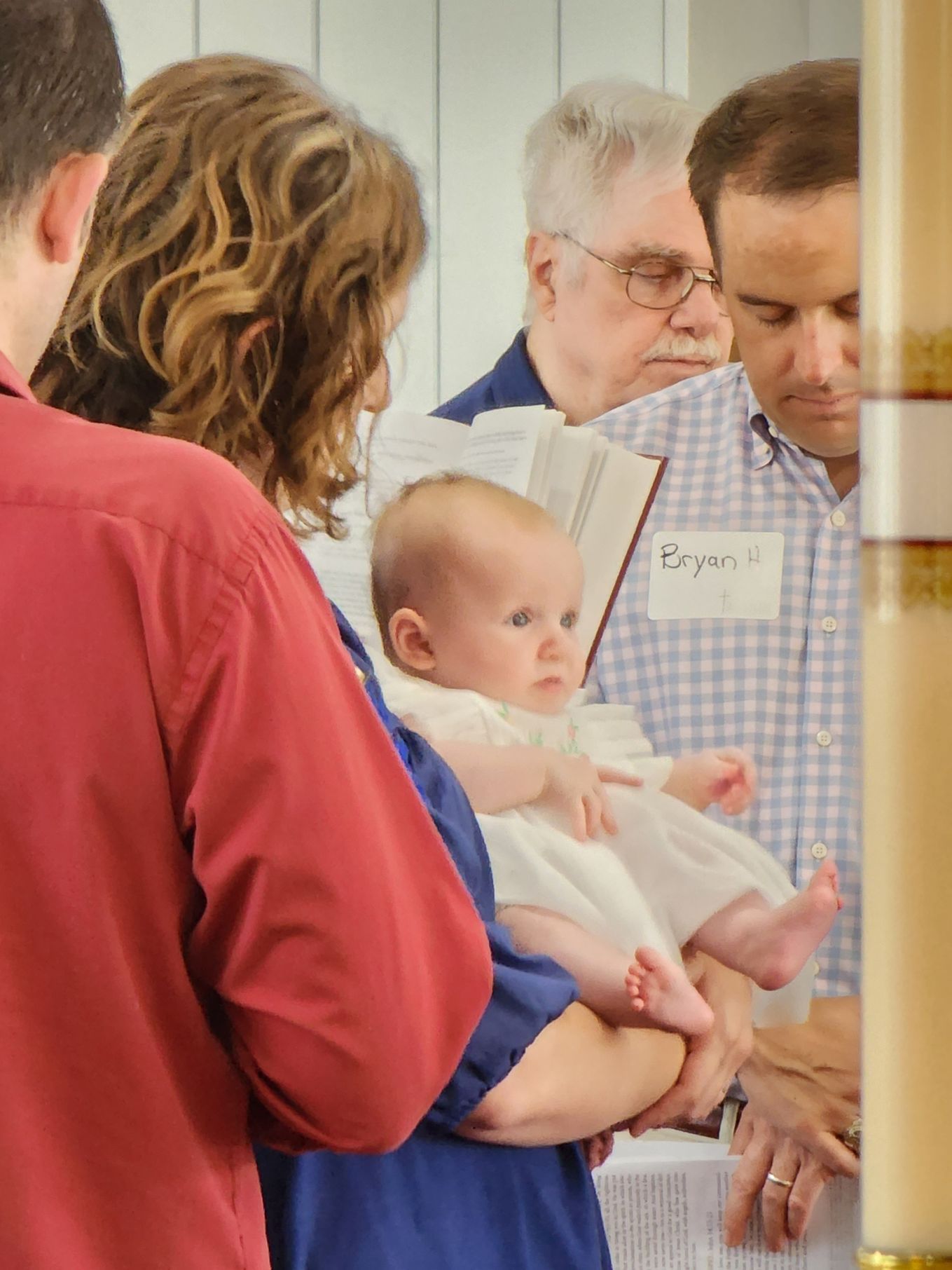 Baby held during a baptism ceremony. People gather around, some reading from a book.