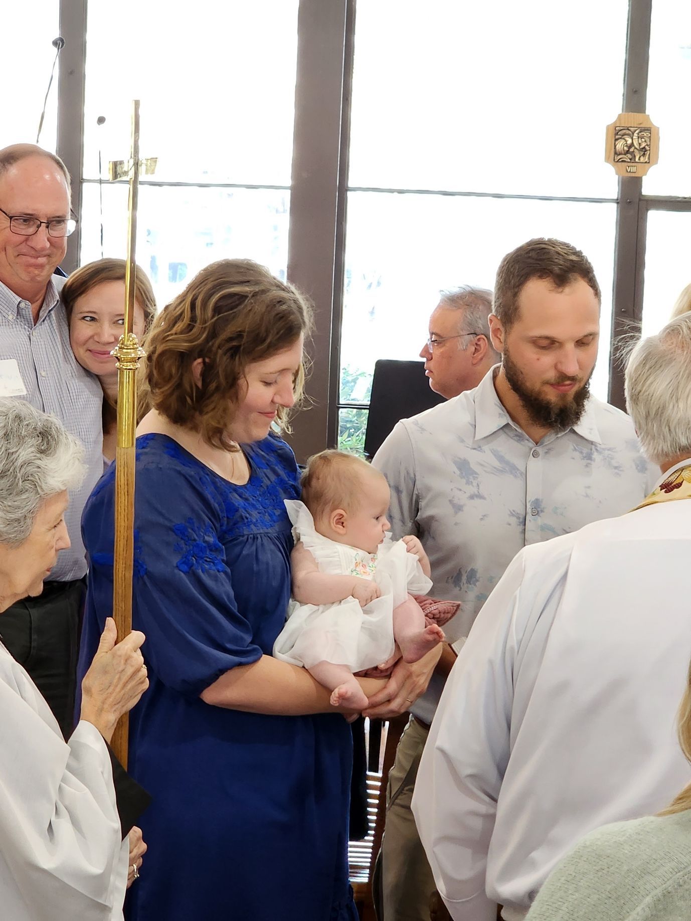 Woman holding baby during baptism ceremony, other attendees in background.