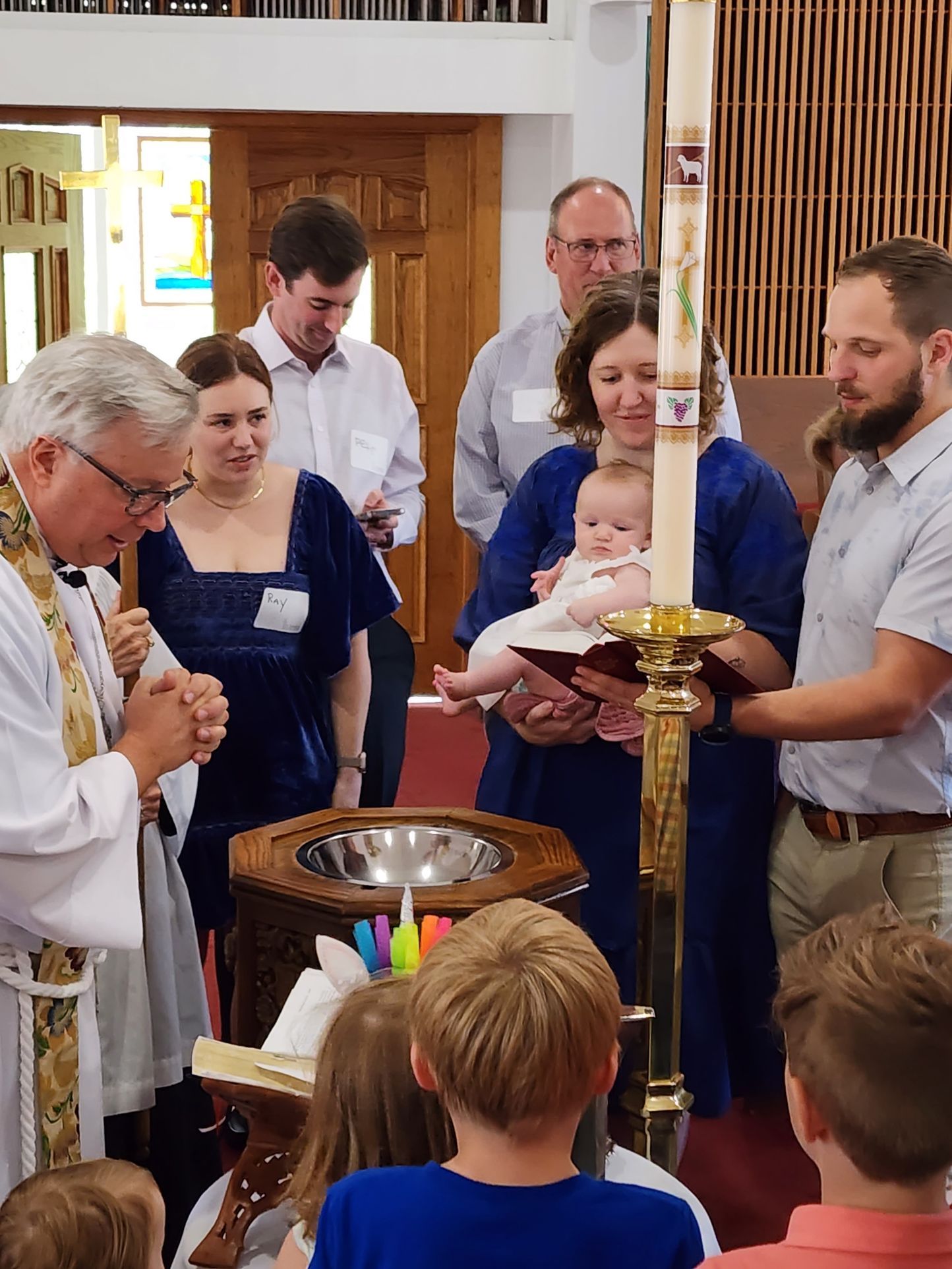 Baptism in a church: priest blesses a baby held by a woman, surrounded by family, with a candle.