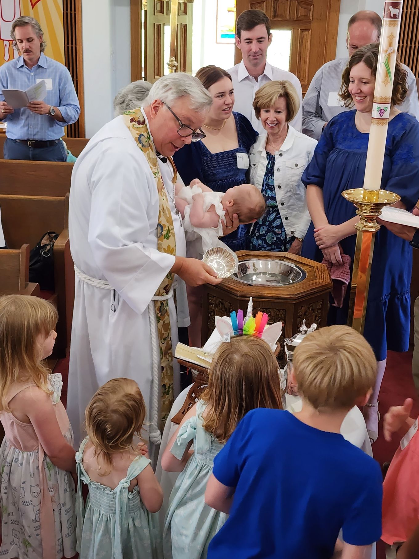 Clergyman baptizes a baby at a church. Several people observe. Children gather in front.