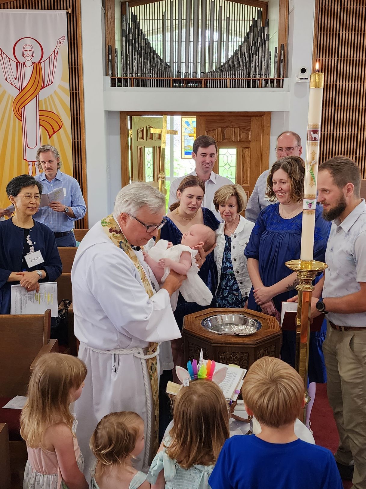 Clergy baptizing a baby in a church, surrounded by family and other attendees.