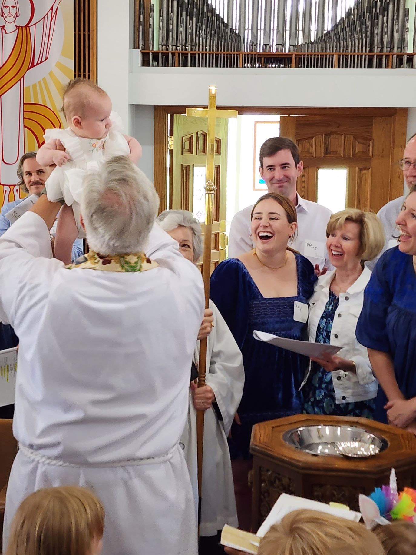 Clergy holding a baby during a baptism ceremony; people smiling, church interior.