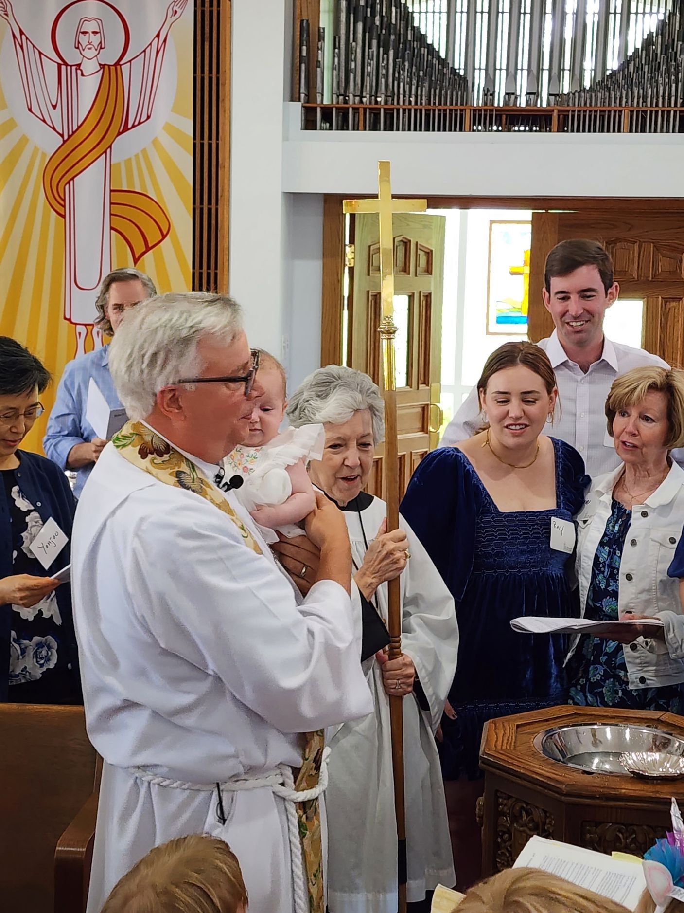 Clergyman holding a baby during a baptism ceremony. Attendees observe near a baptismal font. Bright church interior.