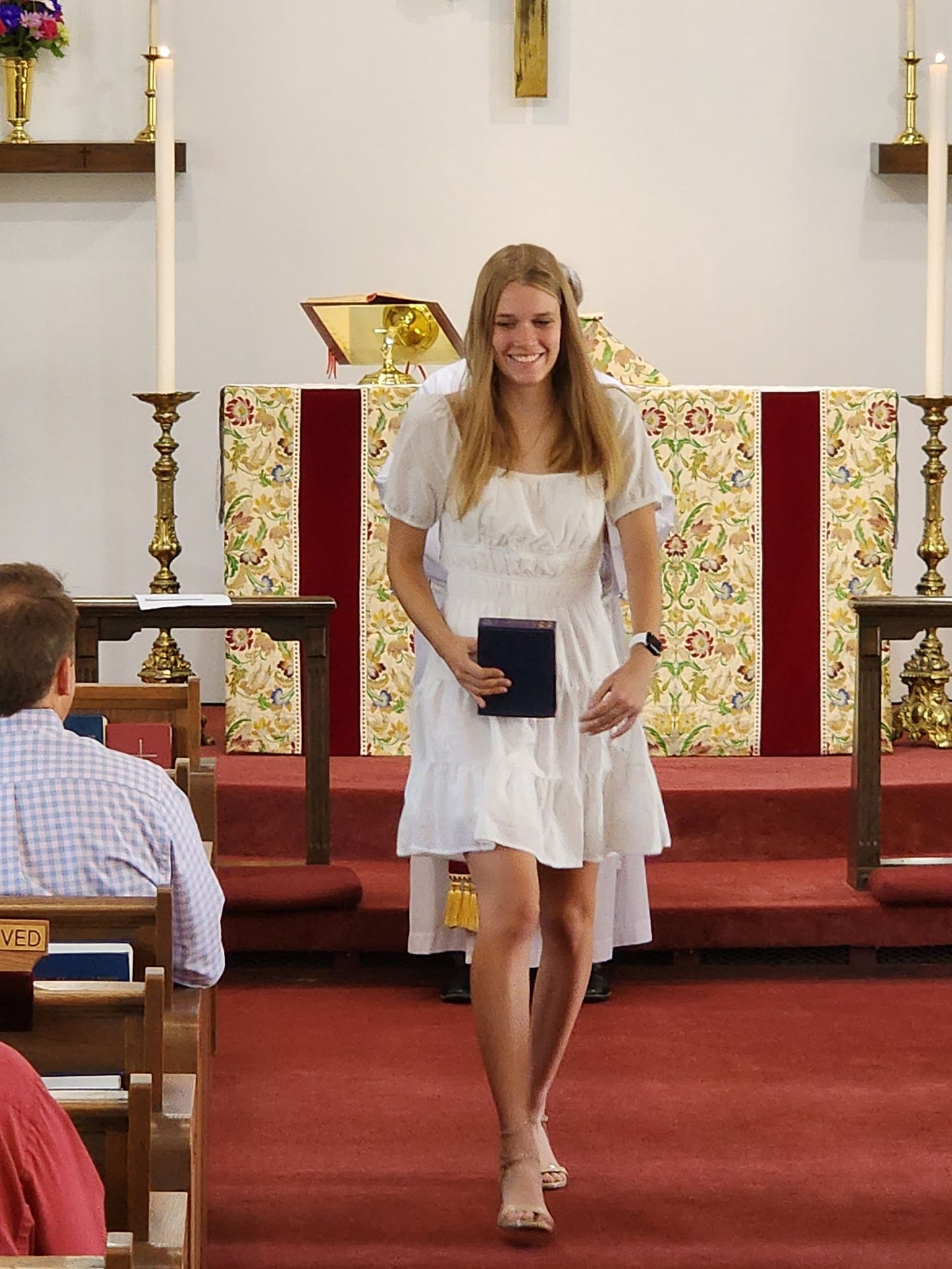 Woman in white dress smiles, holding a book, walking in a church on red carpet.