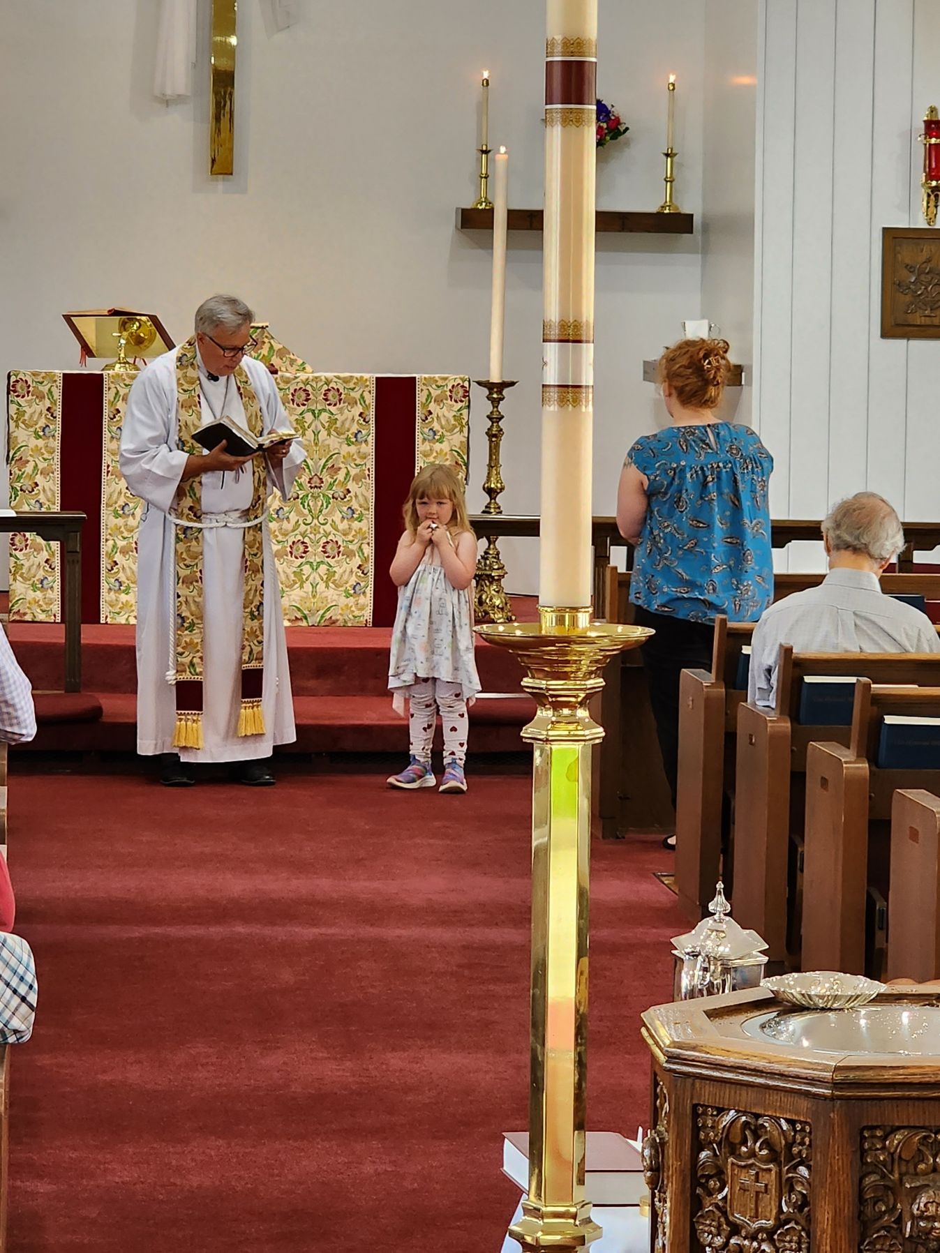 Priest reads to a young girl in a church. A woman and man sit in pews. Gold candle in the foreground.