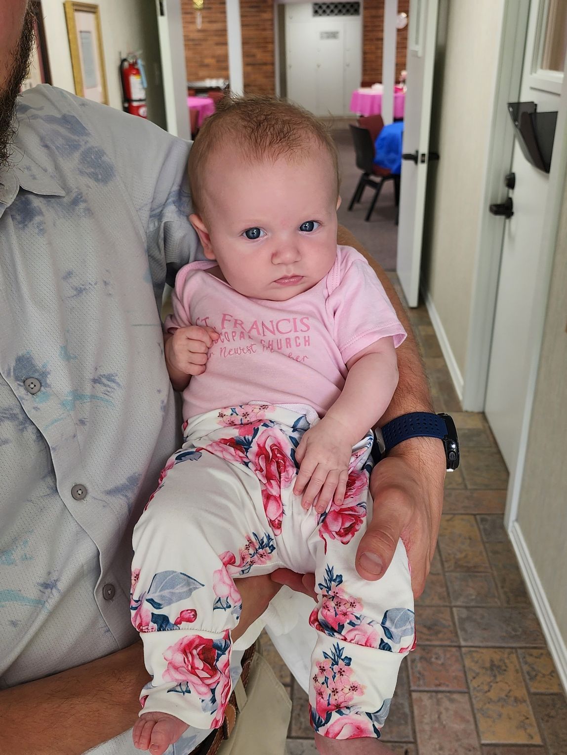 Baby in pink shirt and floral pants held by a person indoors.