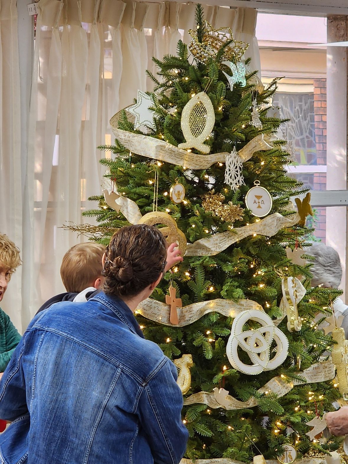 Person and child decorating a Christmas tree with gold ribbon and ornaments.