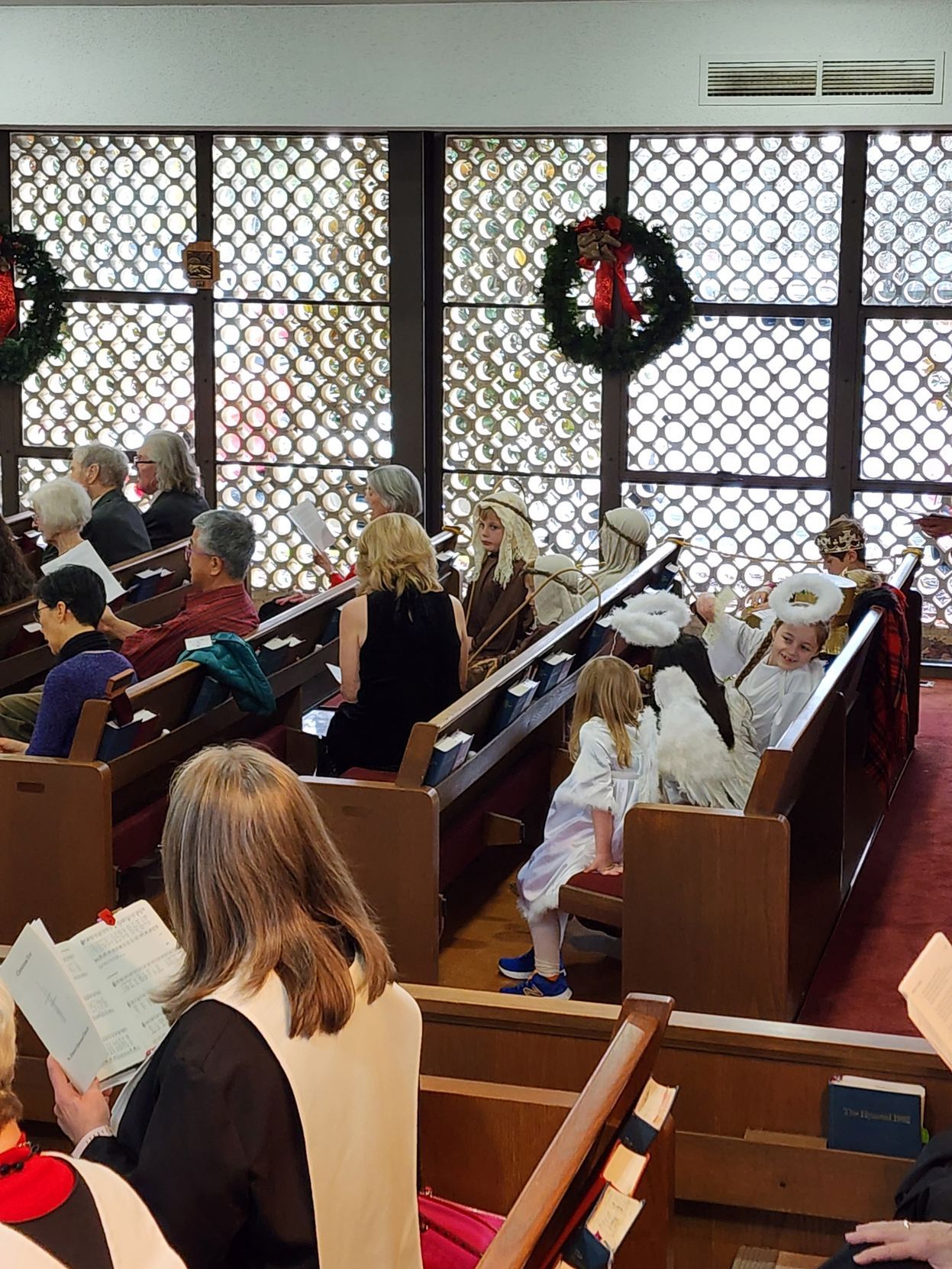 People in church pews, some in costumes, reading and attending a service with Christmas wreaths.