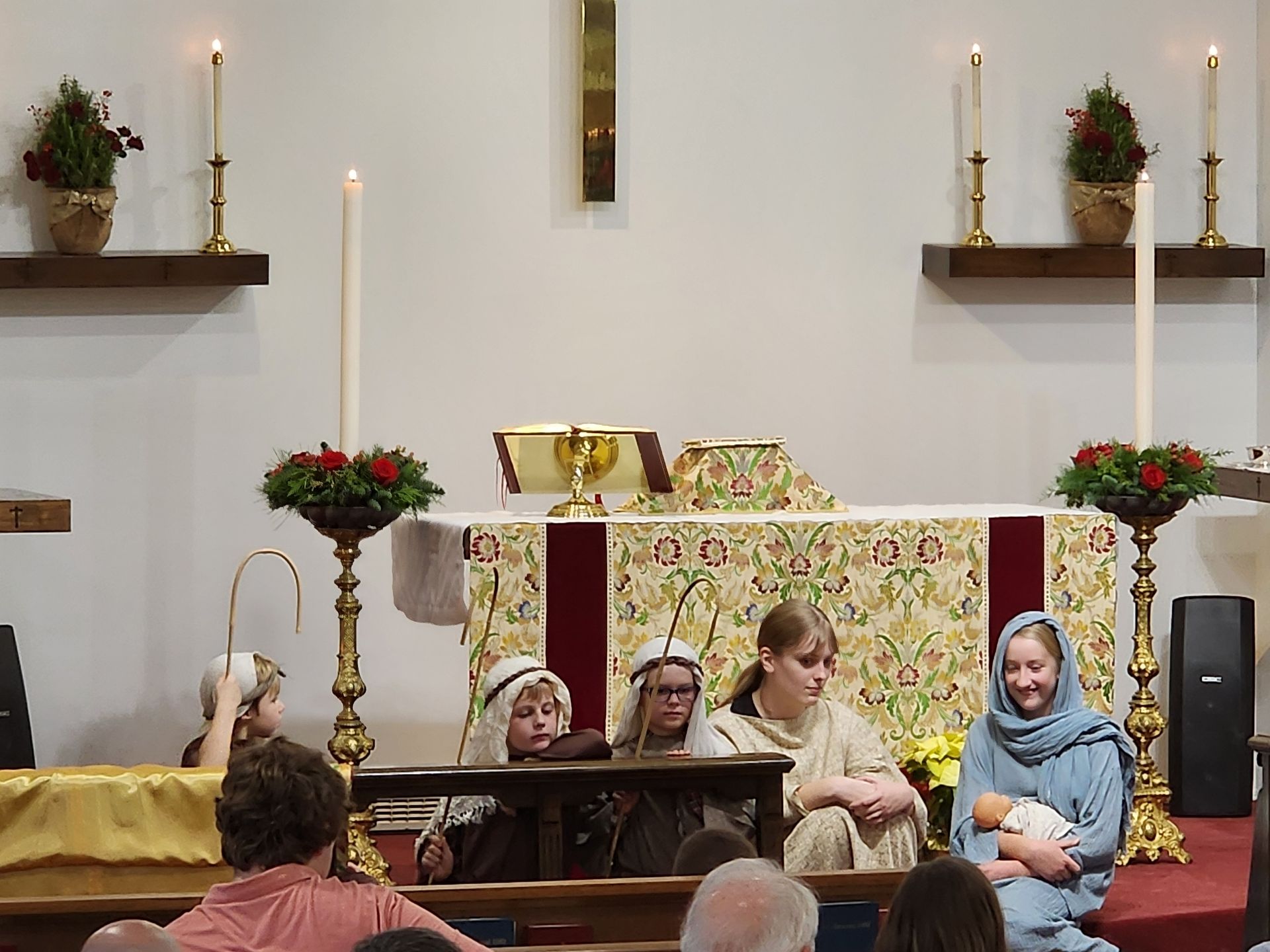 Nativity scene in a church: actors in costumes, holding a baby. Candles, altar, and audience are visible.