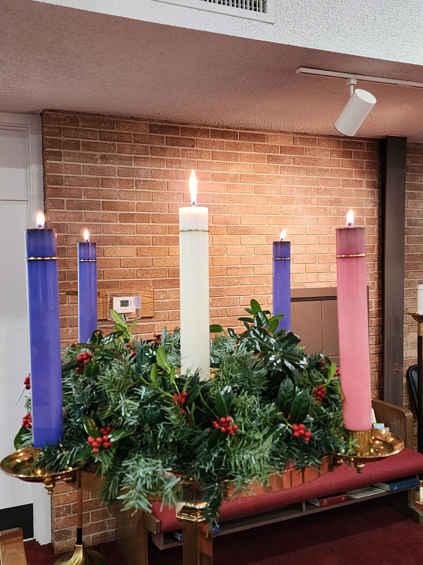 Advent wreath with four lit candles, one unlit, on a table in front of a brick wall.