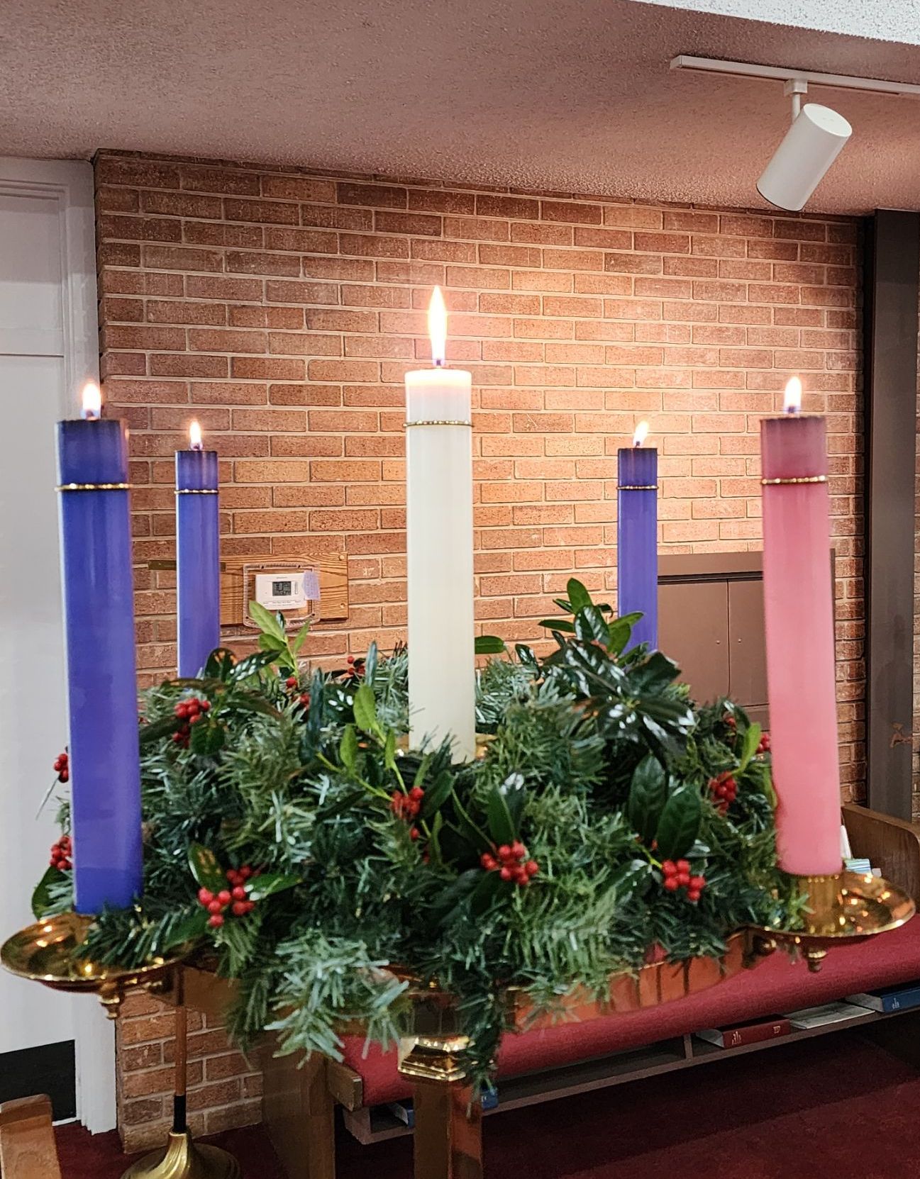 Advent wreath with lit candles in church. Central white candle lit, surrounded by three purple and one pink candle, holly and greenery.