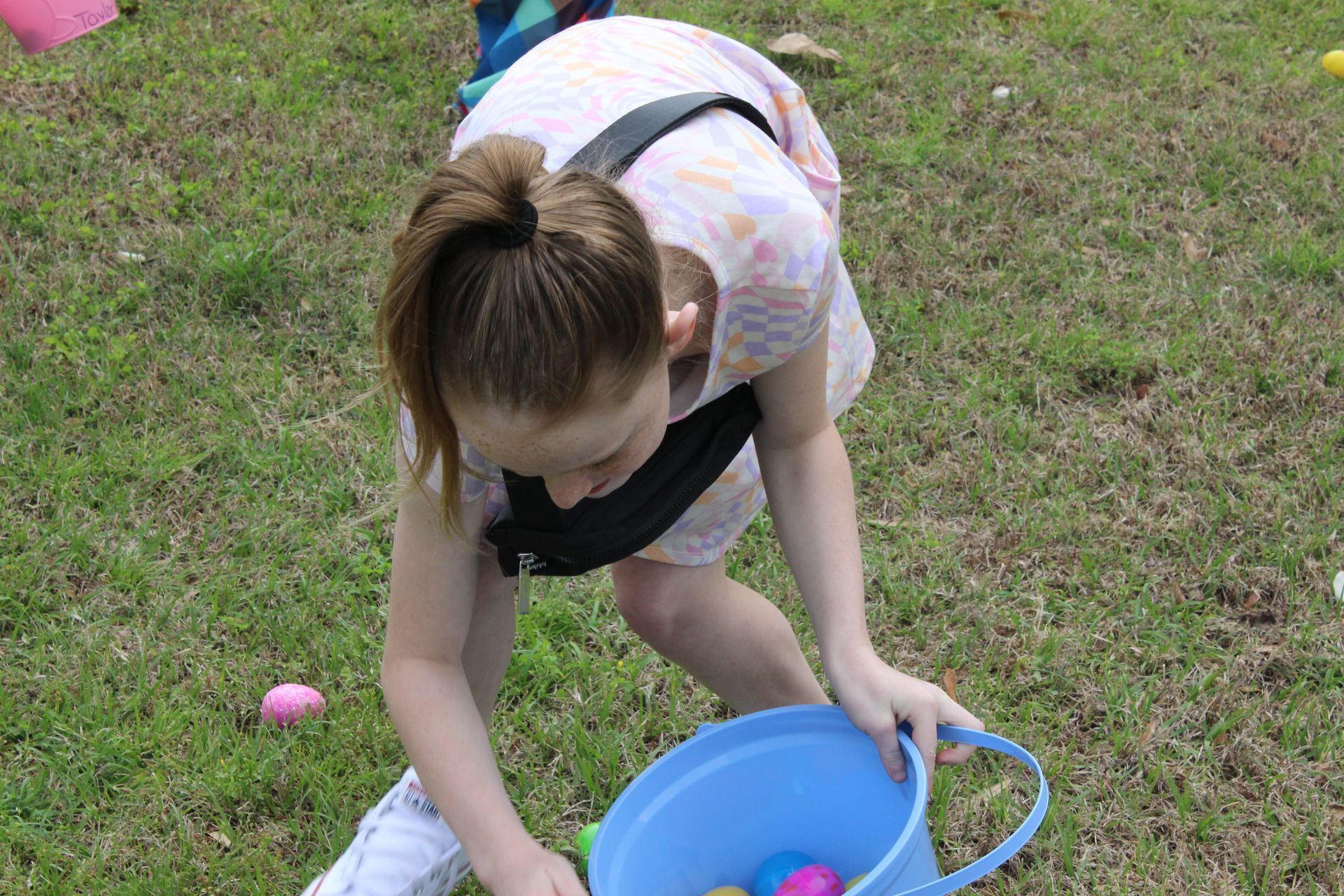 Girl with ponytail collecting Easter eggs from grass into a blue basket.