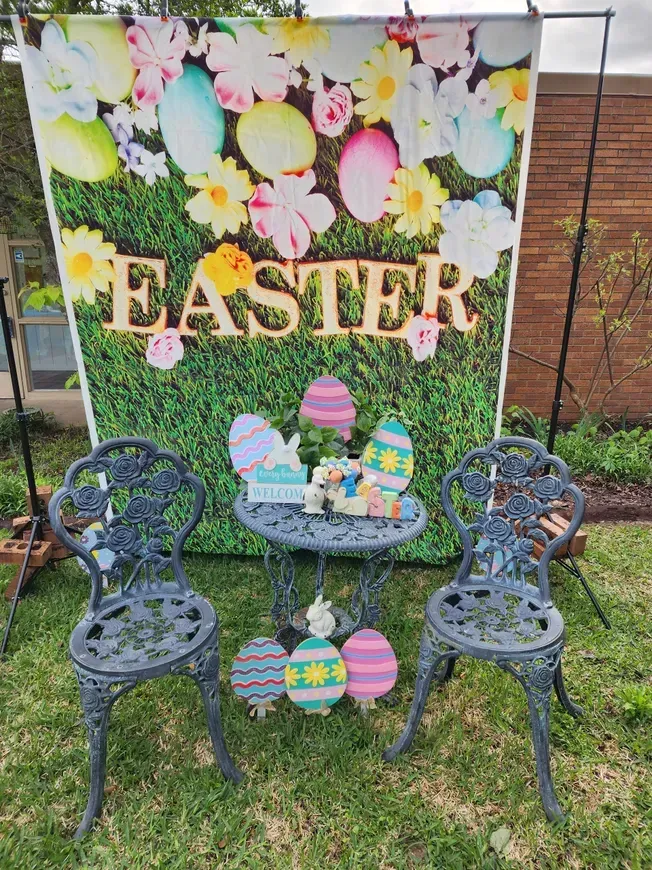 Easter photo booth with backdrop of eggs and flowers, small table, two chairs, decorations.