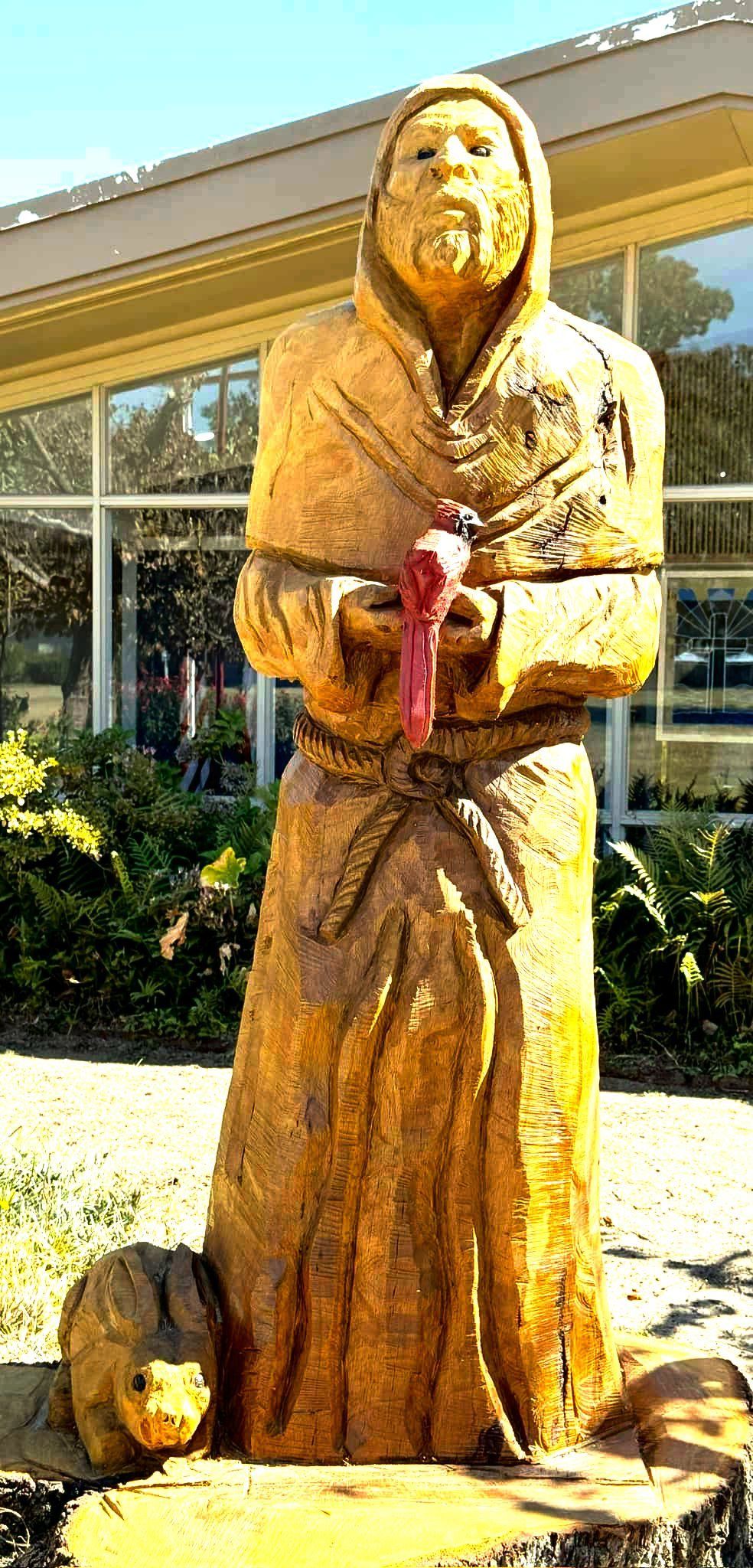 Wooden statue of a robed figure holding a red item, with a small animal at the base, outdoors.