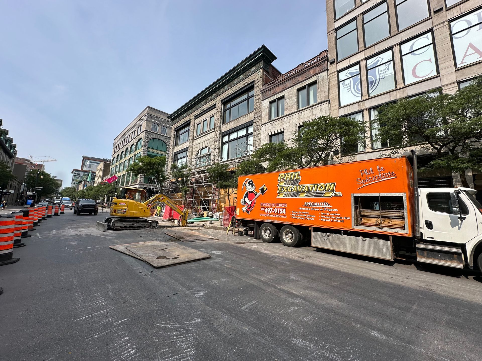 Scène de travaux de voirie avec un camion orange, une excavatrice et des bâtiments.