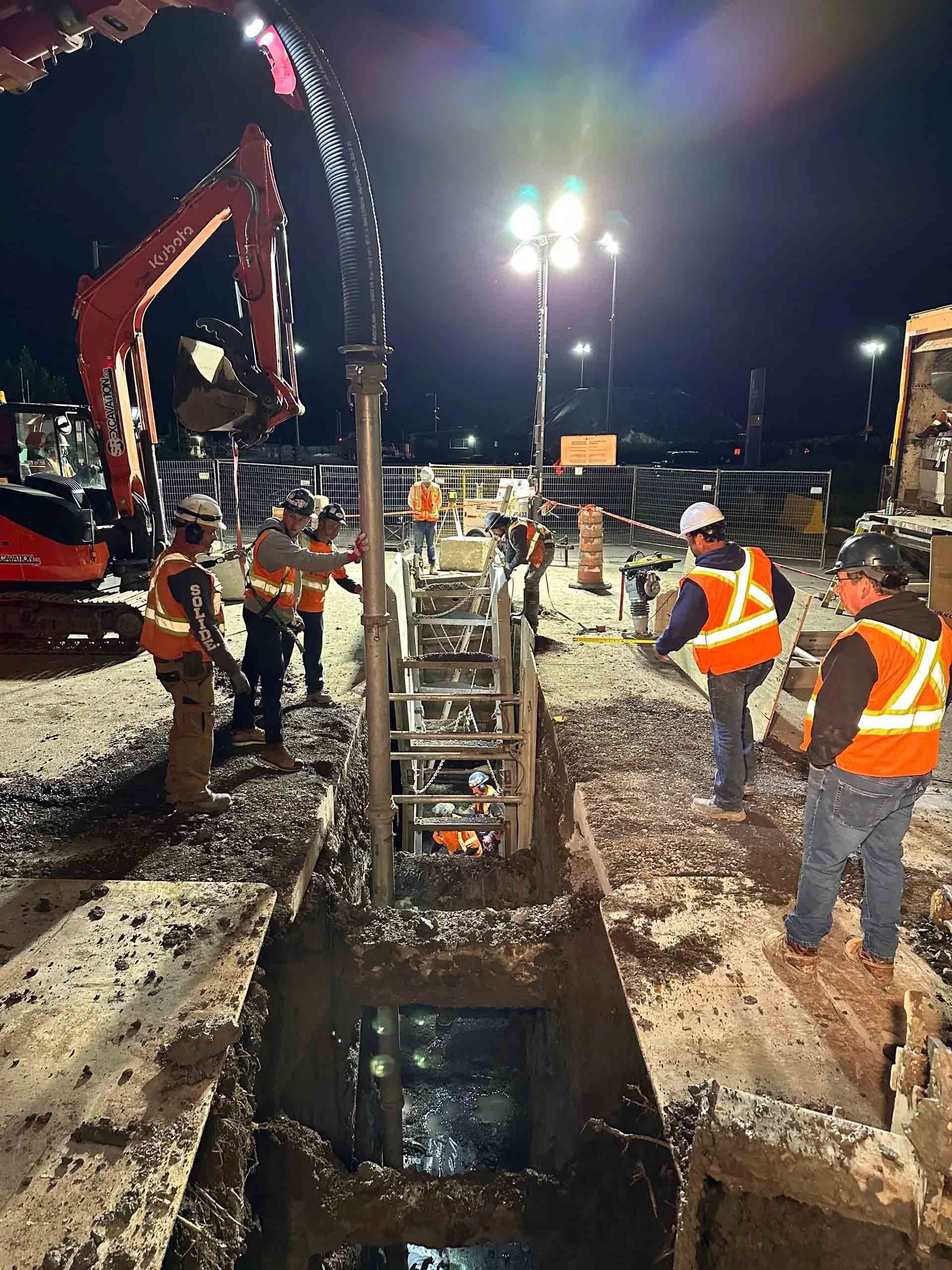 Construction crew working at night, digging in a trench. Several workers in orange vests operate machinery under bright lights.