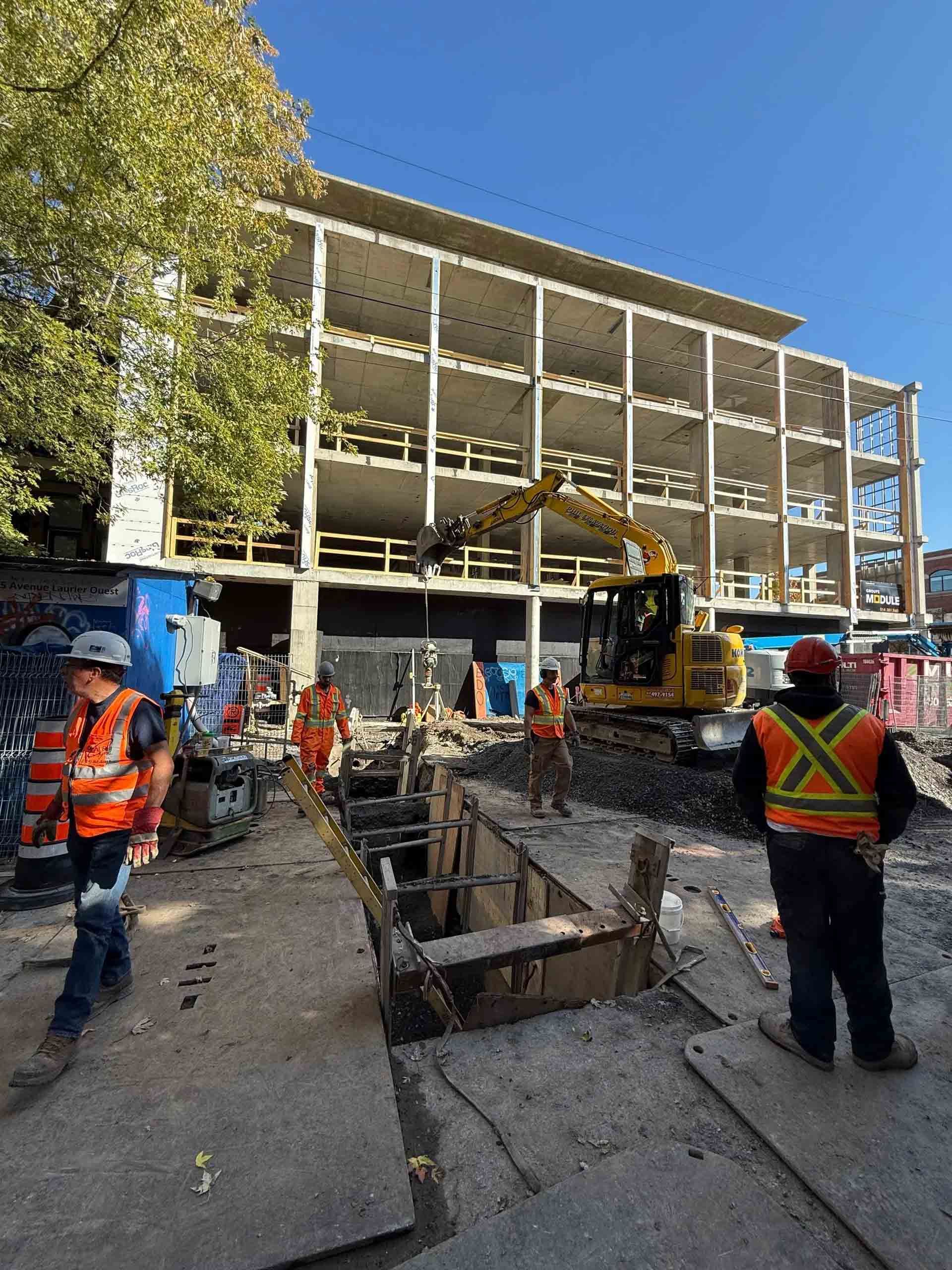 Chantier de construction avec ouvriers, excavatrice et bâtiment en construction sous un ciel bleu.
