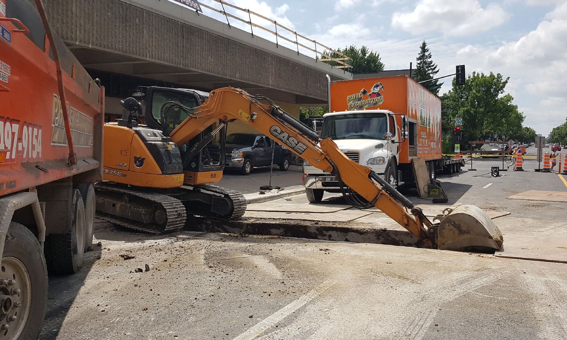 Une excavatrice creuse dans une rue sous un pont ; des camions de chantier et des cônes de signalisation sont présents.